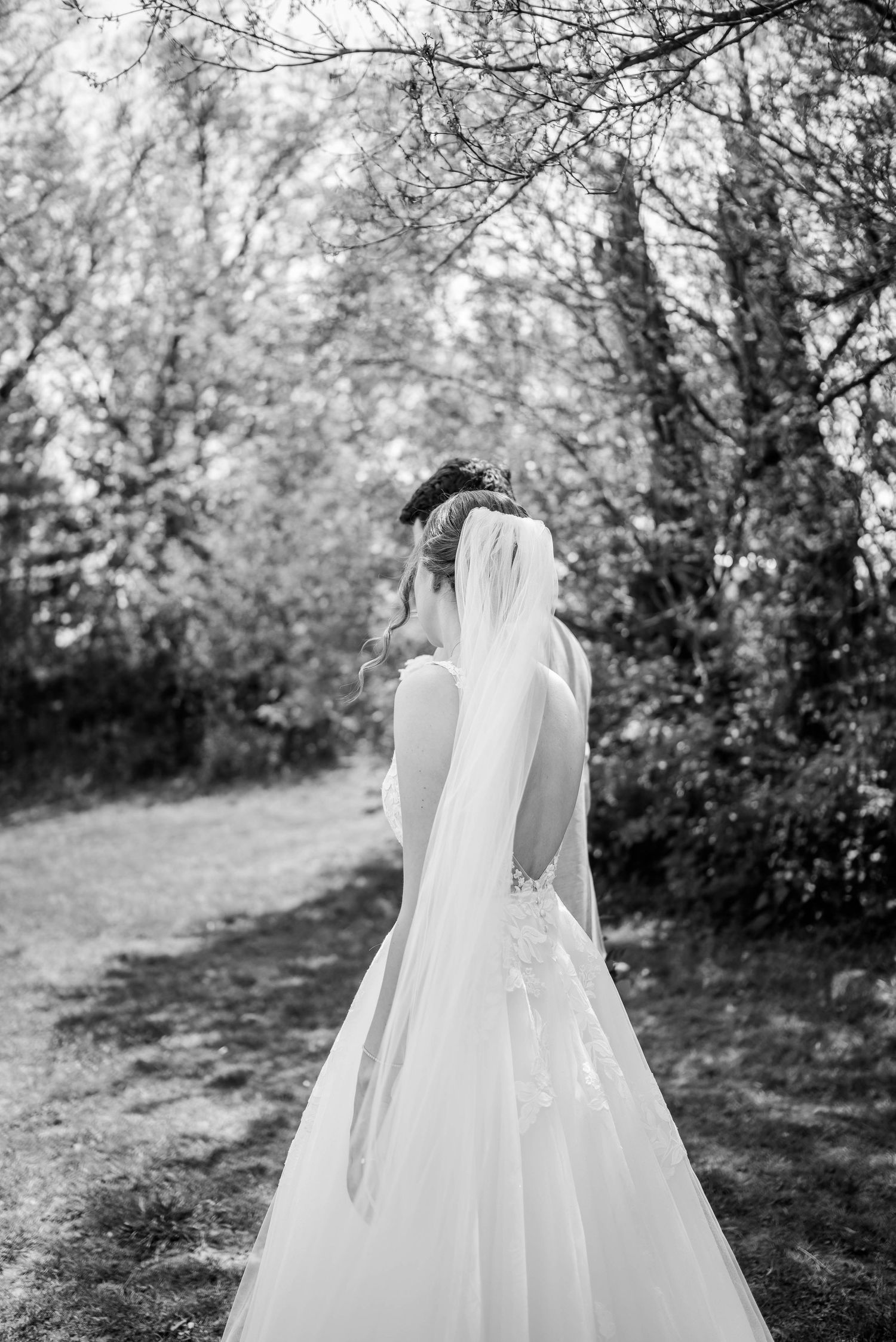 Black and white portrait of bride in backless gown and veil walking through wooded path.