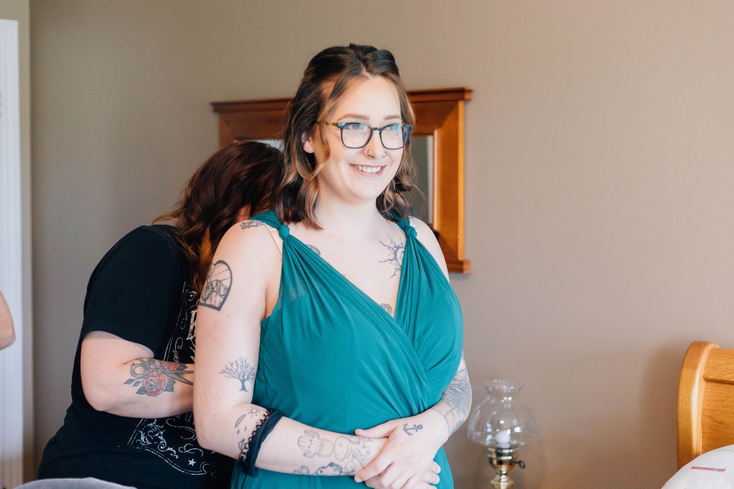 Smiling woman with glasses in teal dress being helped by friend in bedroom.