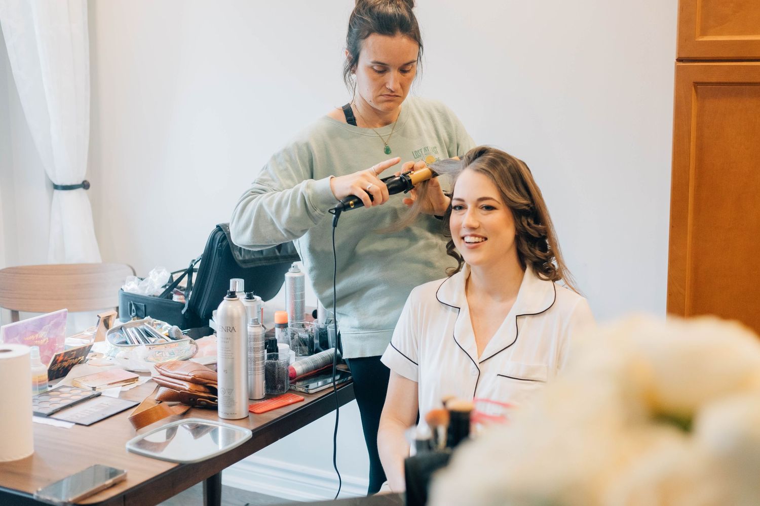 Hair stylist curling bride's hair while she smiles during wedding morning preparations.