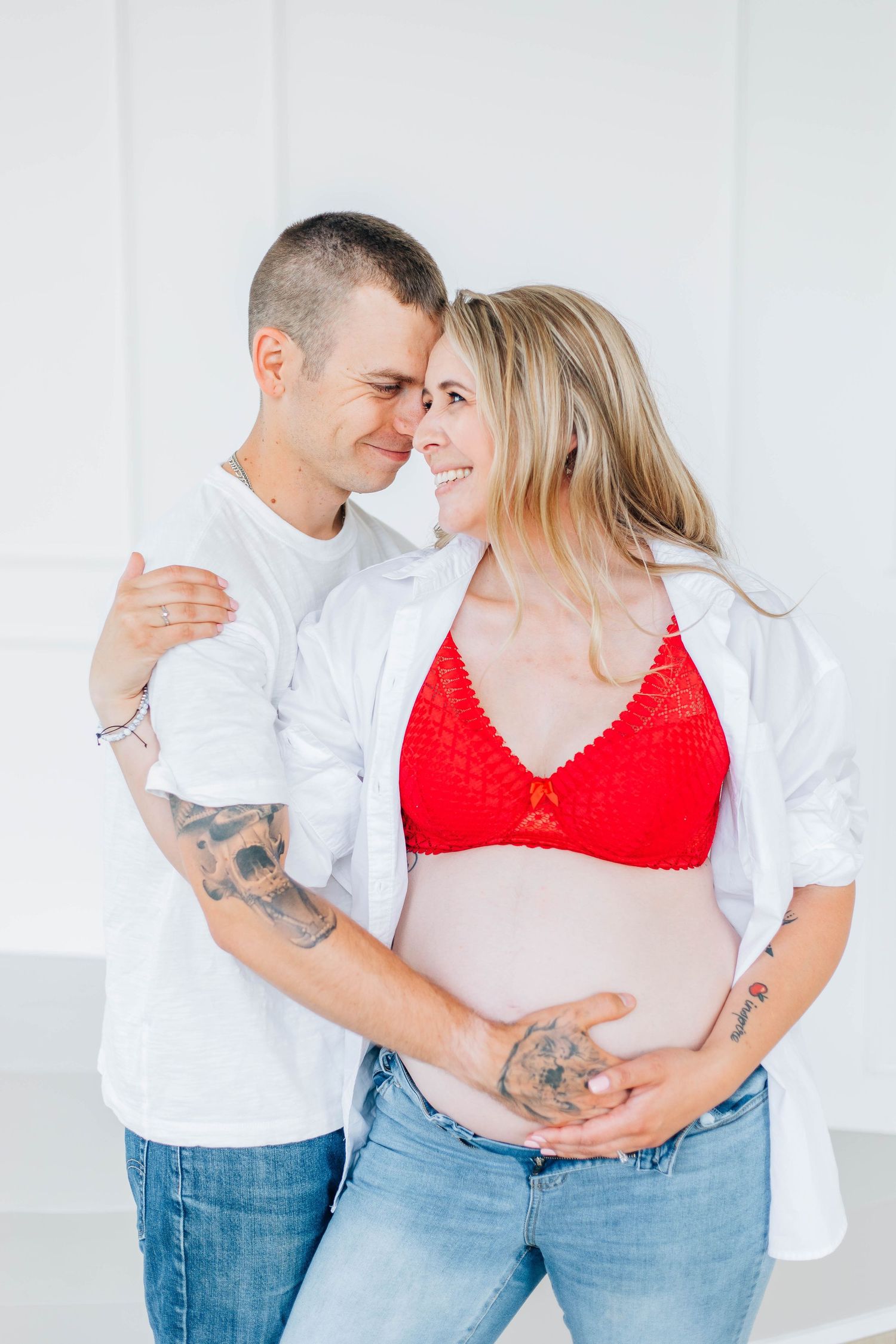 Tattooed couple smiling together while cradling pregnant belly in white studio.