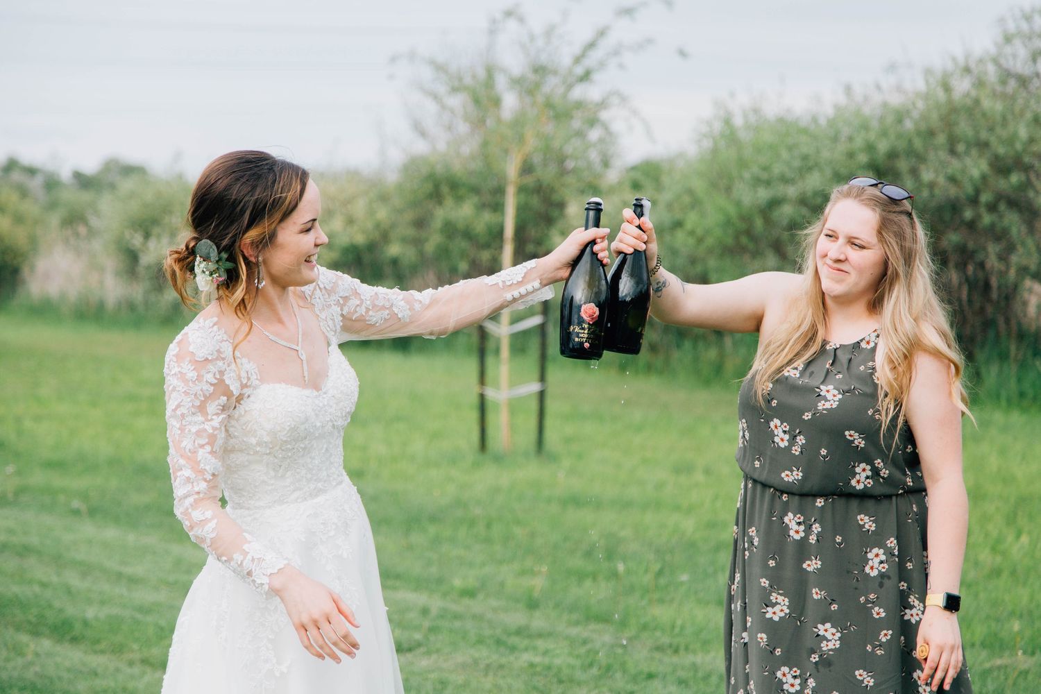 Bride and bridesmaid toasting champagne bottles together on a lush green lawn.