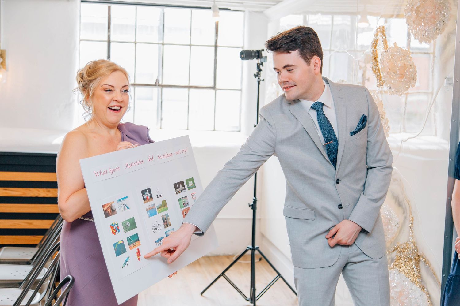 Laughing bridesmaid in lavender dress shows colorful wedding poster to smiling groom at indoor venue.