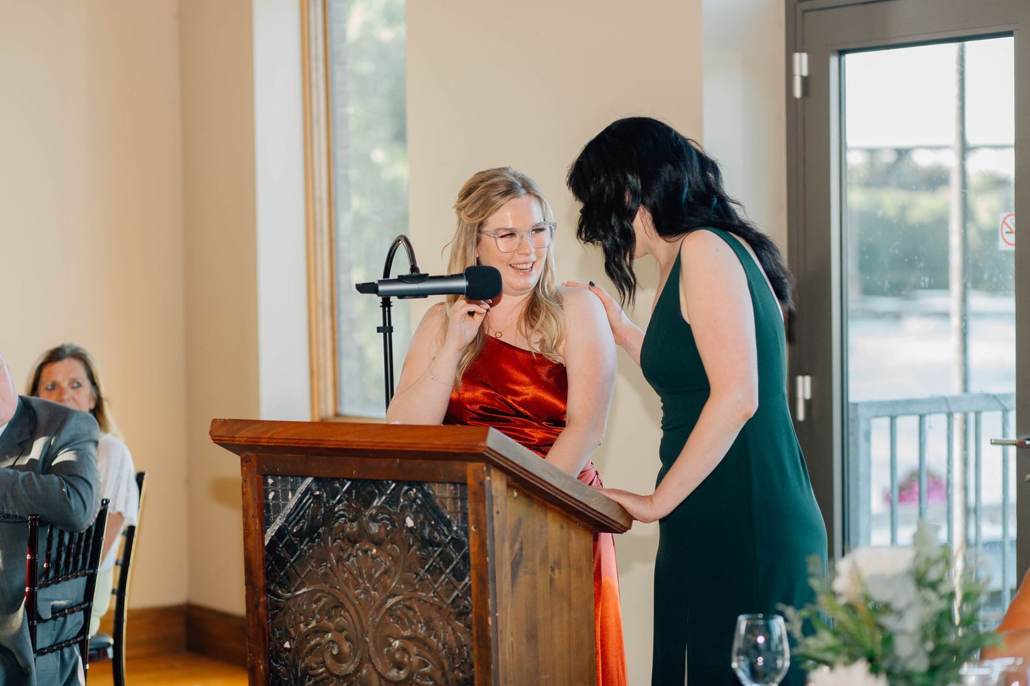 Two bridesmaids in red and green dresses deliver a heartfelt speech at a wooden podium during wedding reception.