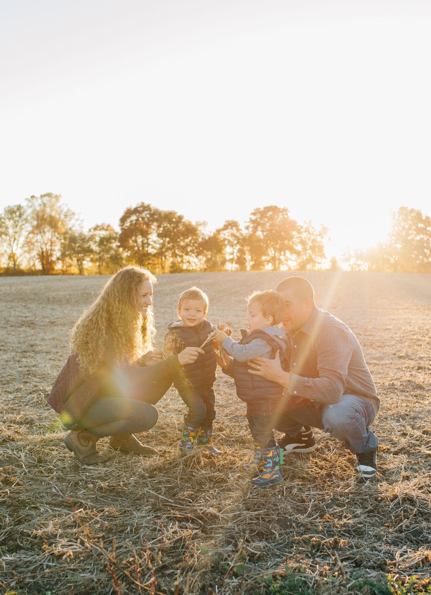 Young family of four crouching together in a golden sunlit field at sunset.