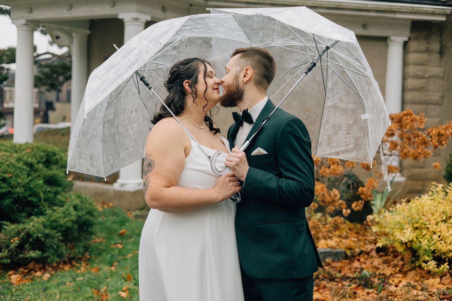 Bride and groom kissing under a clear umbrella surrounded by colorful autumn foliage outdoors.