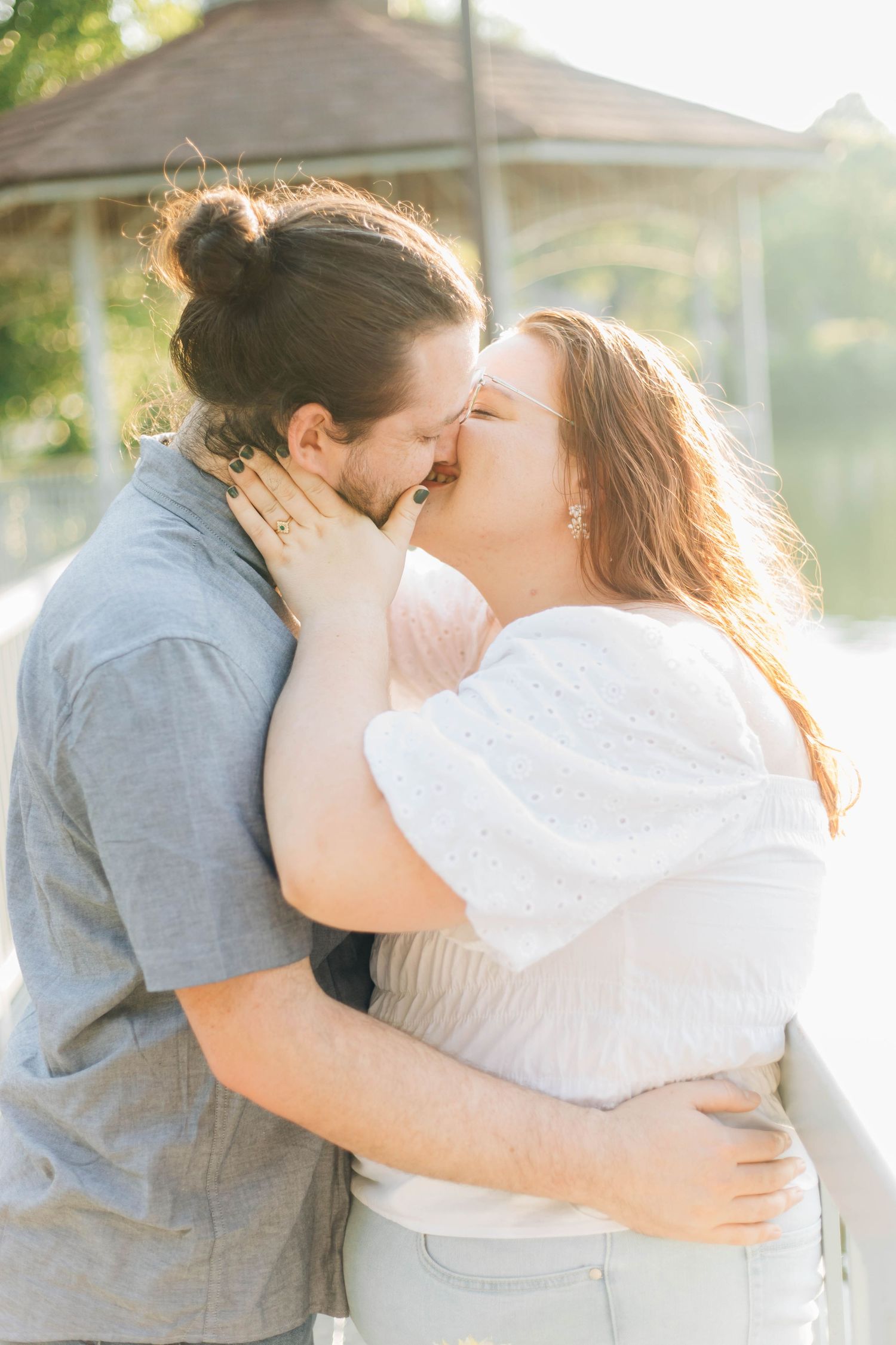 Couple sharing a passionate kiss outdoors in warm golden sunlight near a lakeside gazebo.