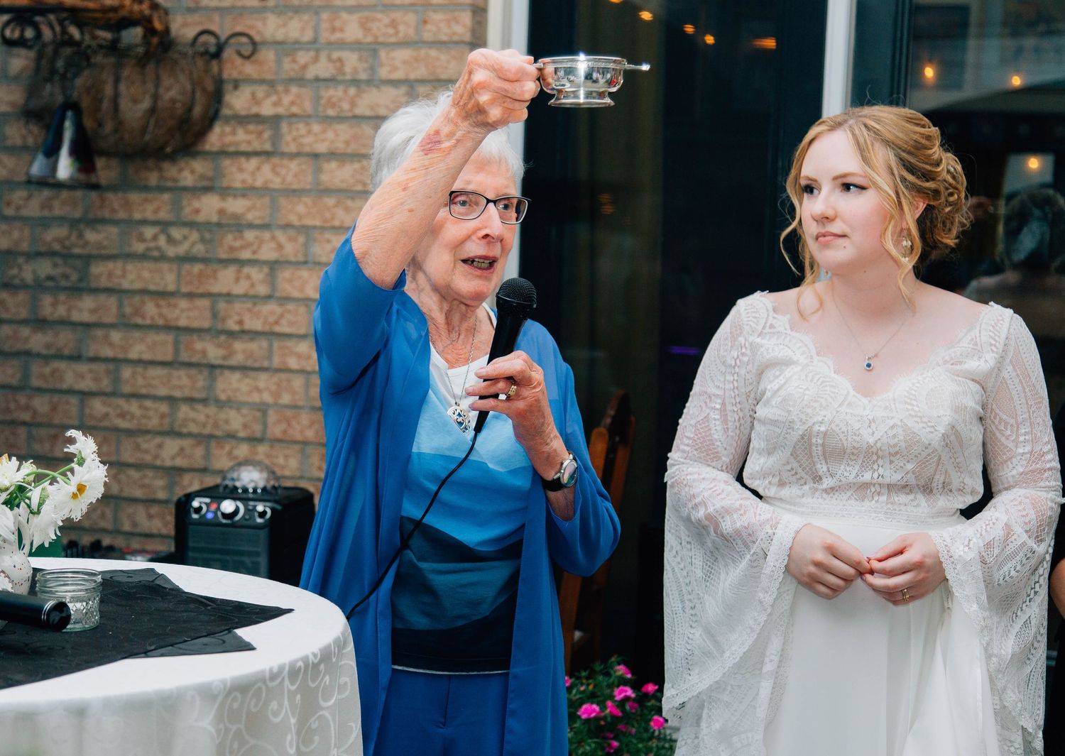 Elderly woman raises a teacup in a toast beside a smiling bride at an outdoor wedding.