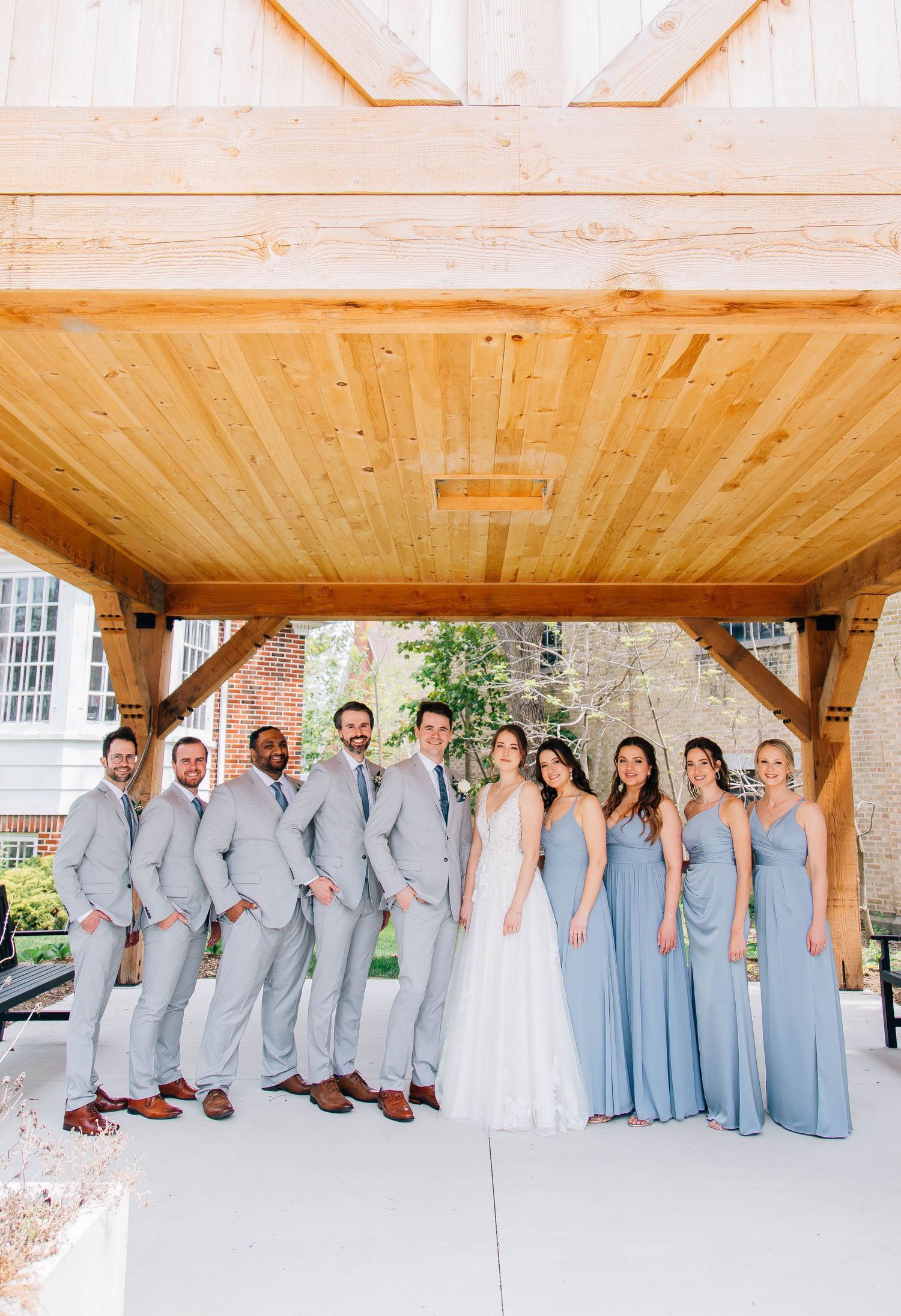 Wedding party poses under wooden pavilion with groomsmen in gray suits and bridesmaids in blue dresses.