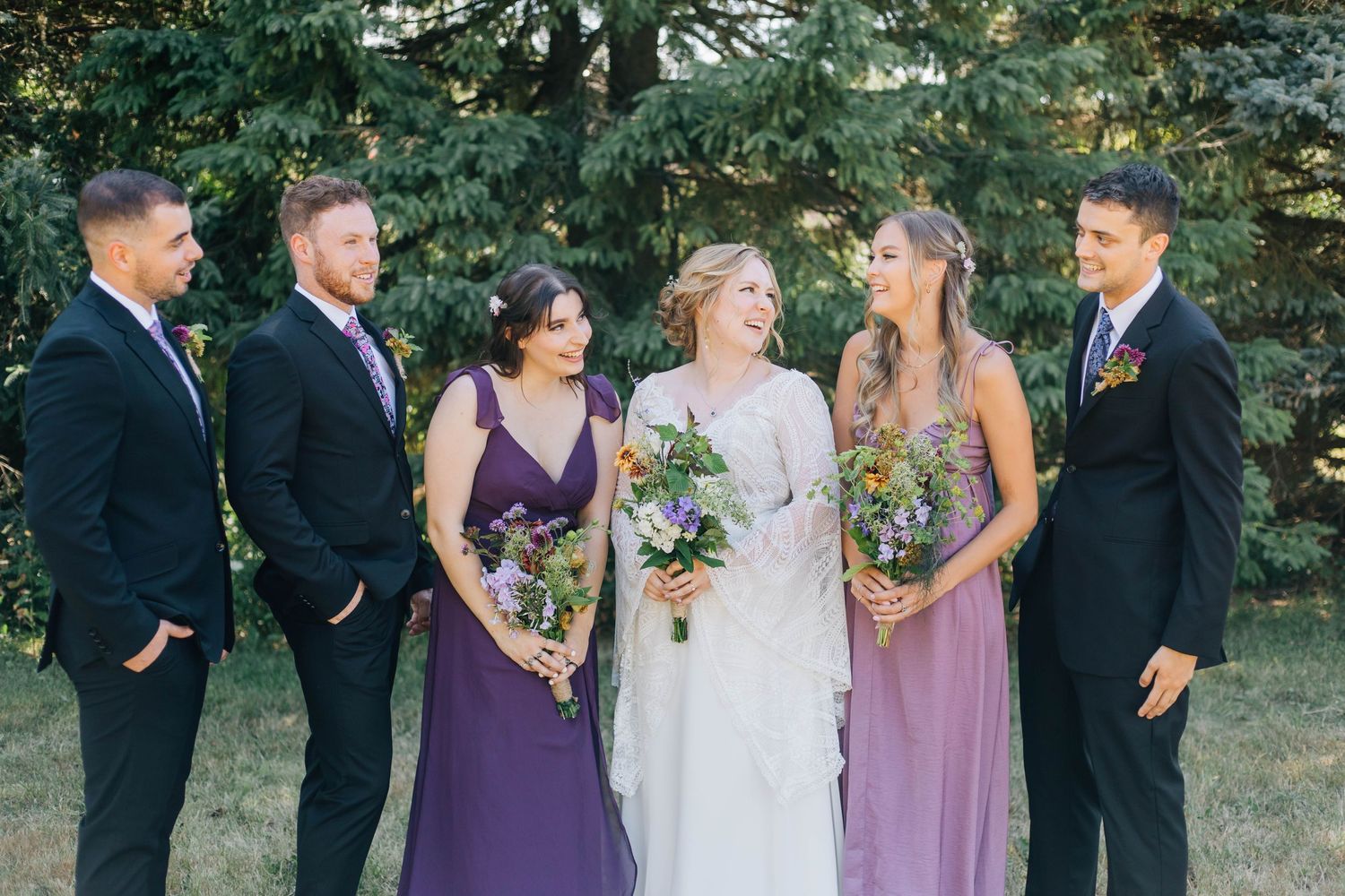 Bride in white gown laughing with bridesmaids in purple dresses and groomsmen in black suits outdoors.