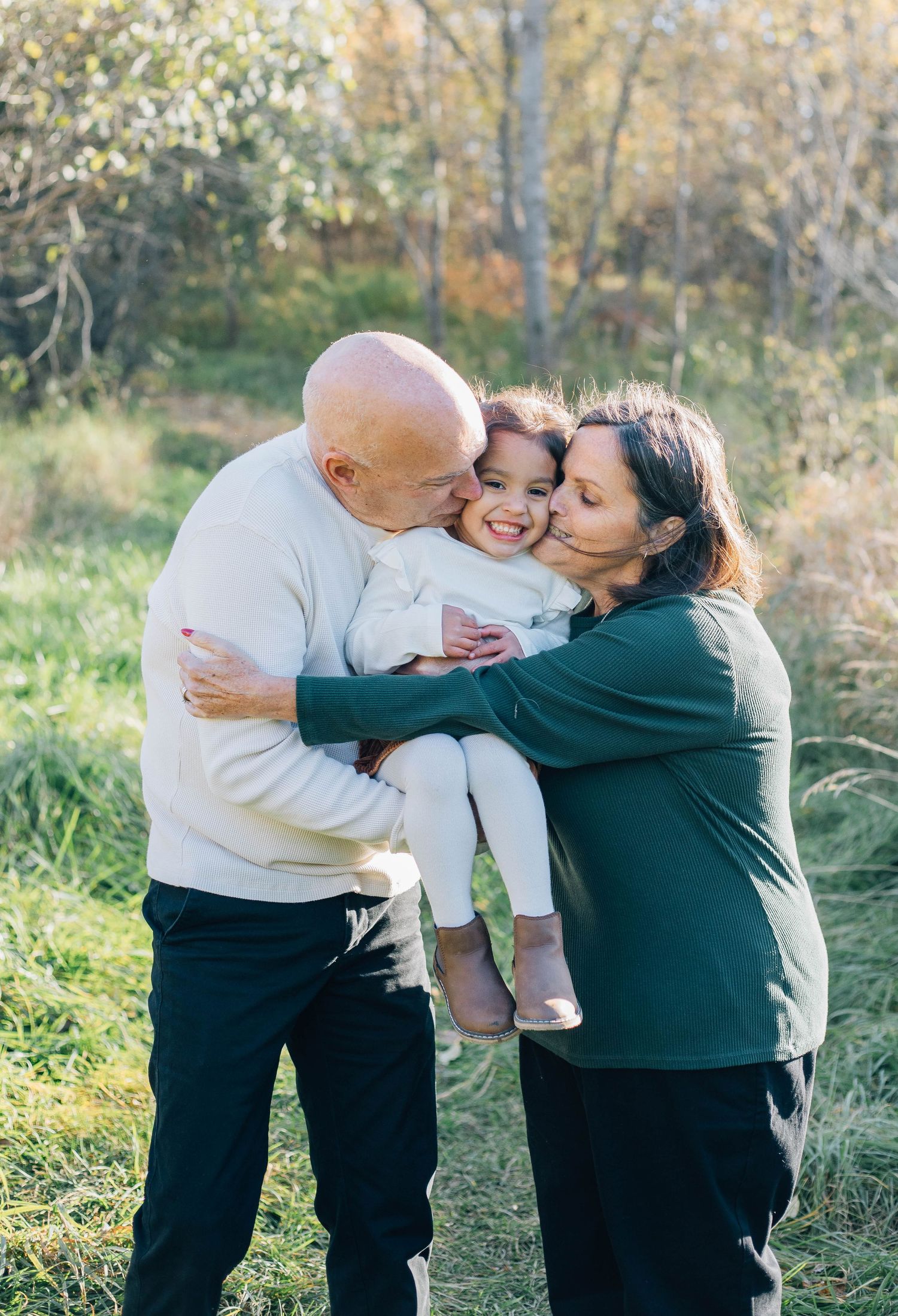 Grandparents kissing and hugging their smiling granddaughter outdoors in a sunny natural setting.