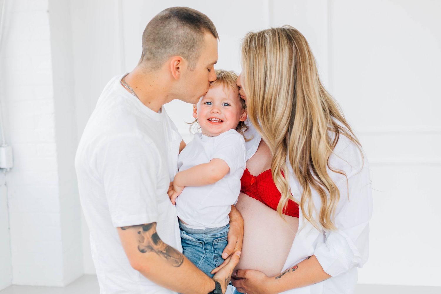 Parents sharing a kiss on cheeks of happy toddler during bright white studio maternity photo session.