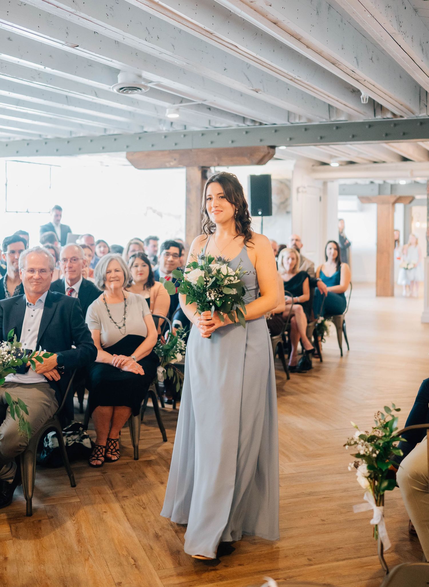 Bridesmaid in grey gown walking down aisle holding bouquet at wedding.