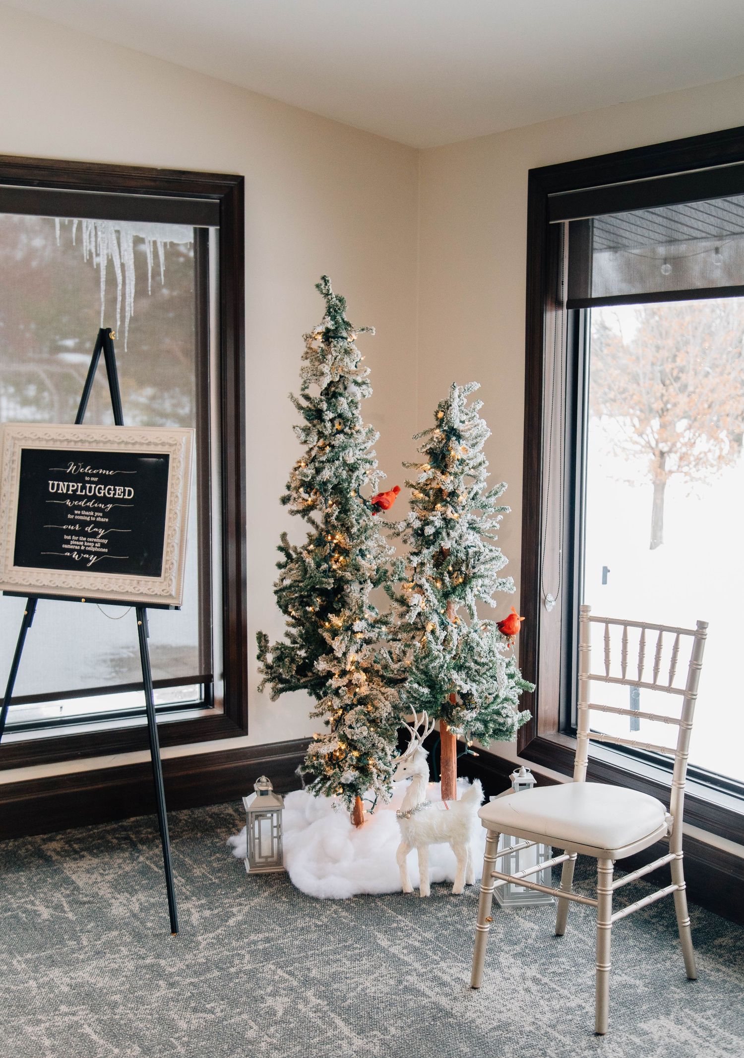 Winter wedding decor featuring flocked Christmas trees with reindeer figurines and an unplugged ceremony sign on an easel.