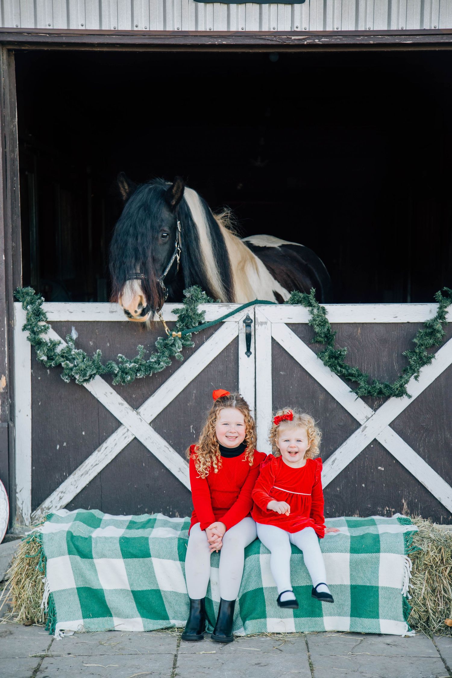 Two young girls in red dresses sit on hay bales in front of a barn stall with a horse.