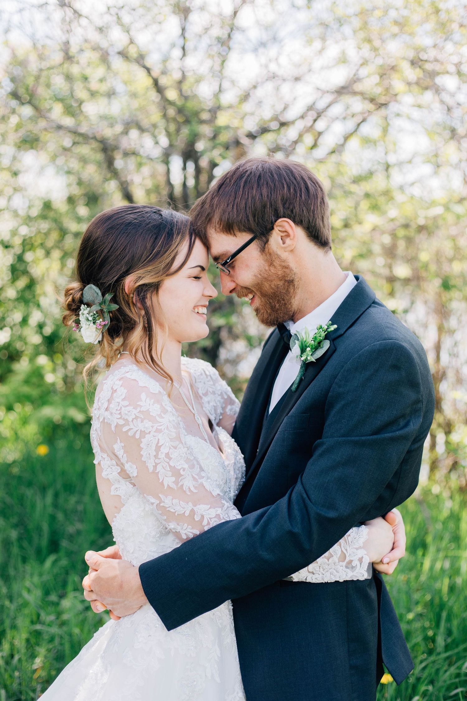 Smiling bride and groom touching foreheads tenderly, embracing in a blooming green outdoor setting.