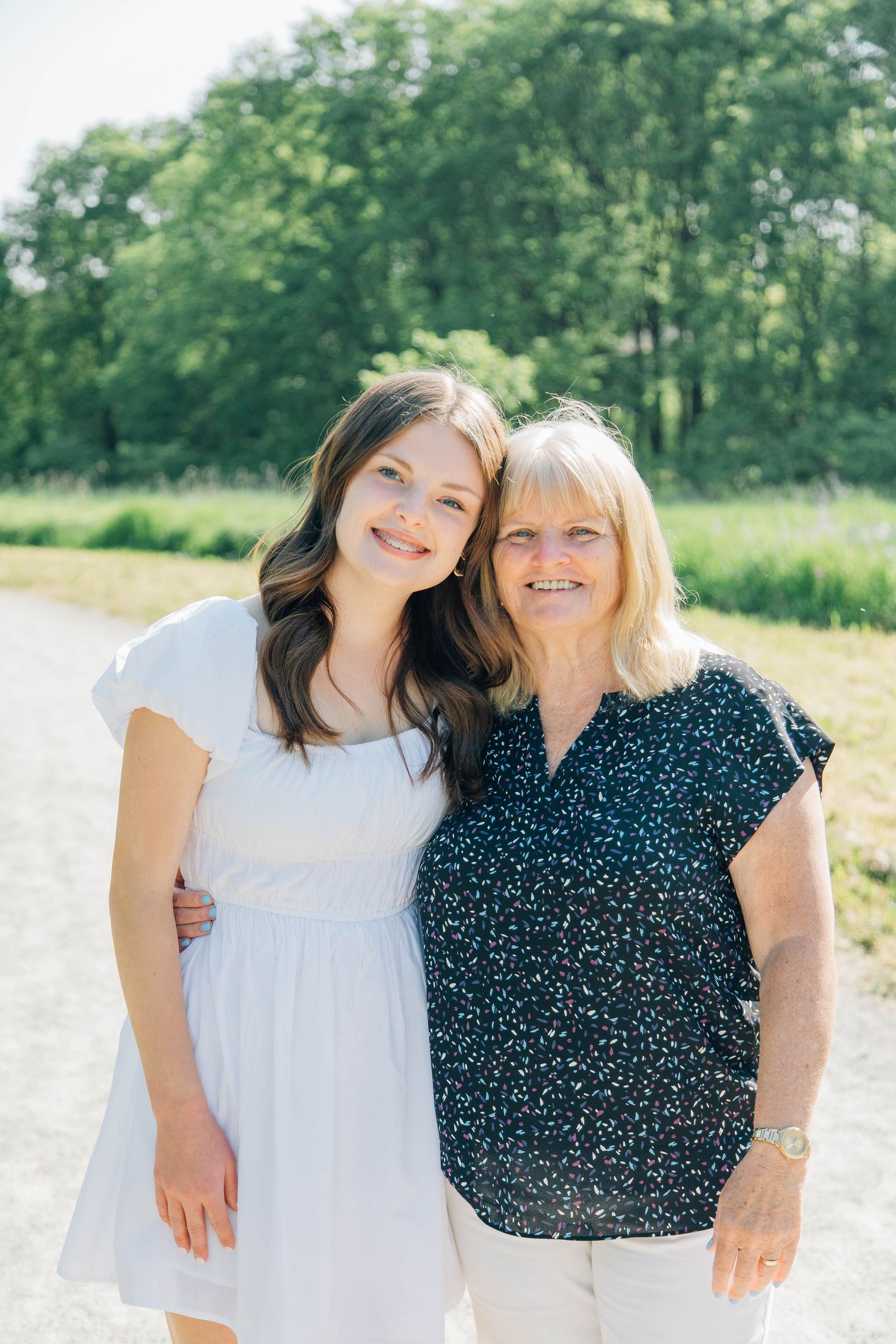 A young woman in a white dress and an older blonde woman smiling together on a sunny outdoor path.