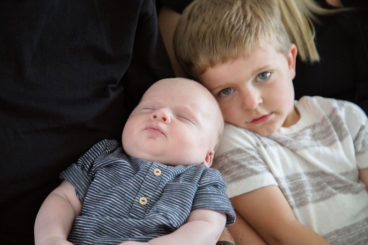 Close-up of young boy tenderly resting cheek against sleeping newborn sibling in blue striped onesie.