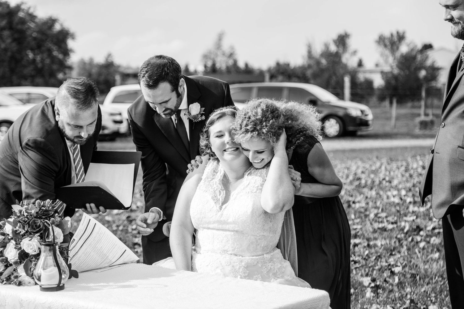 Bride laughs emotionally while signing wedding documents surrounded by wedding party in black and white.