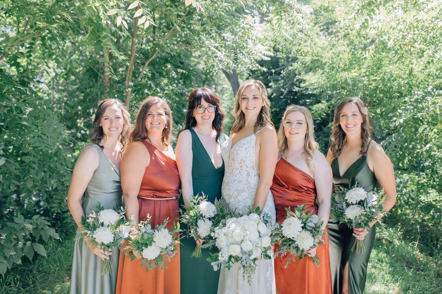 Bride with five bridesmaids in mixed earth-tone dresses holding white floral bouquets.