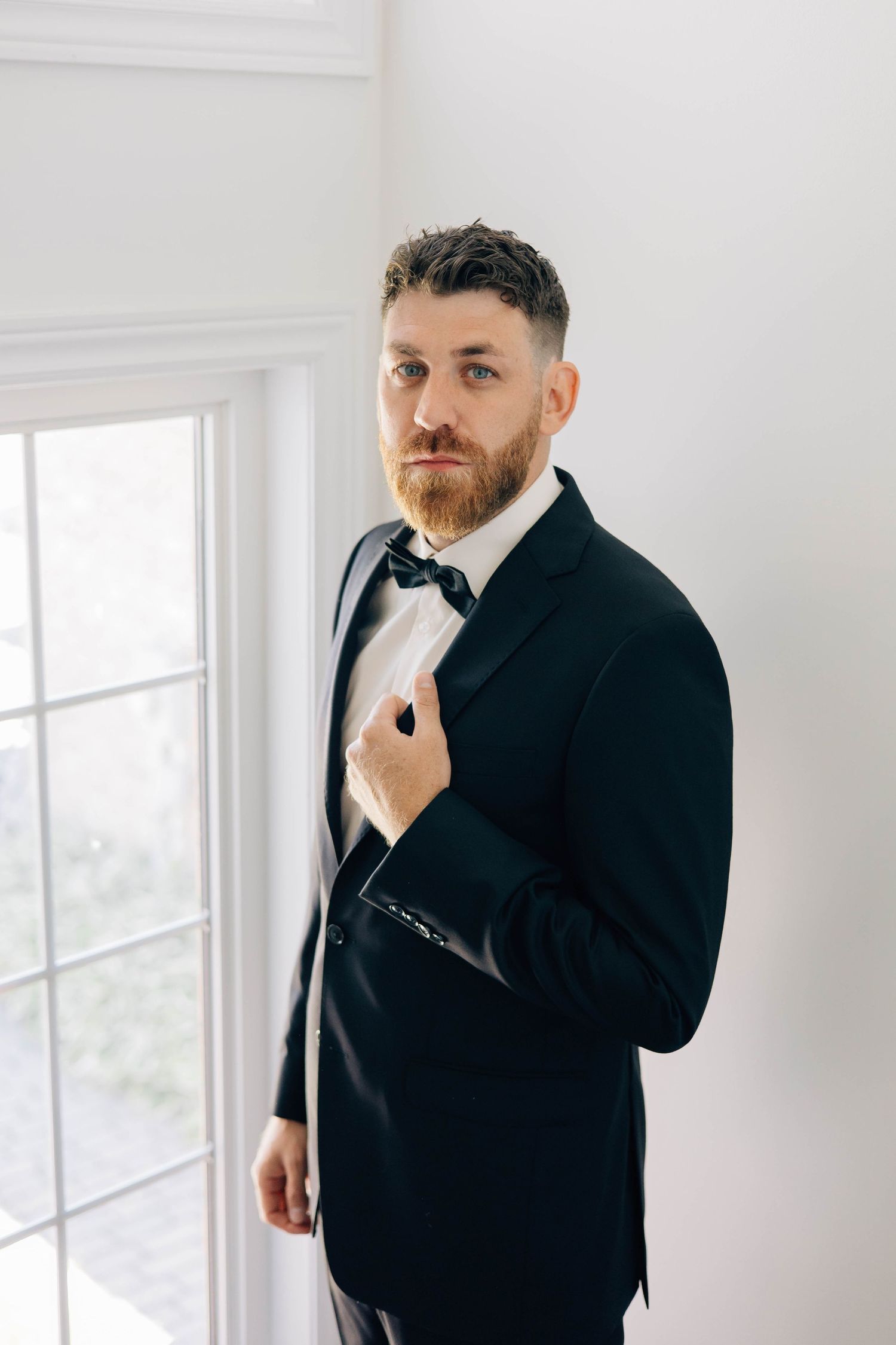 Handsome groom in a black tuxedo and bow tie standing by a bright window before his wedding ceremony.