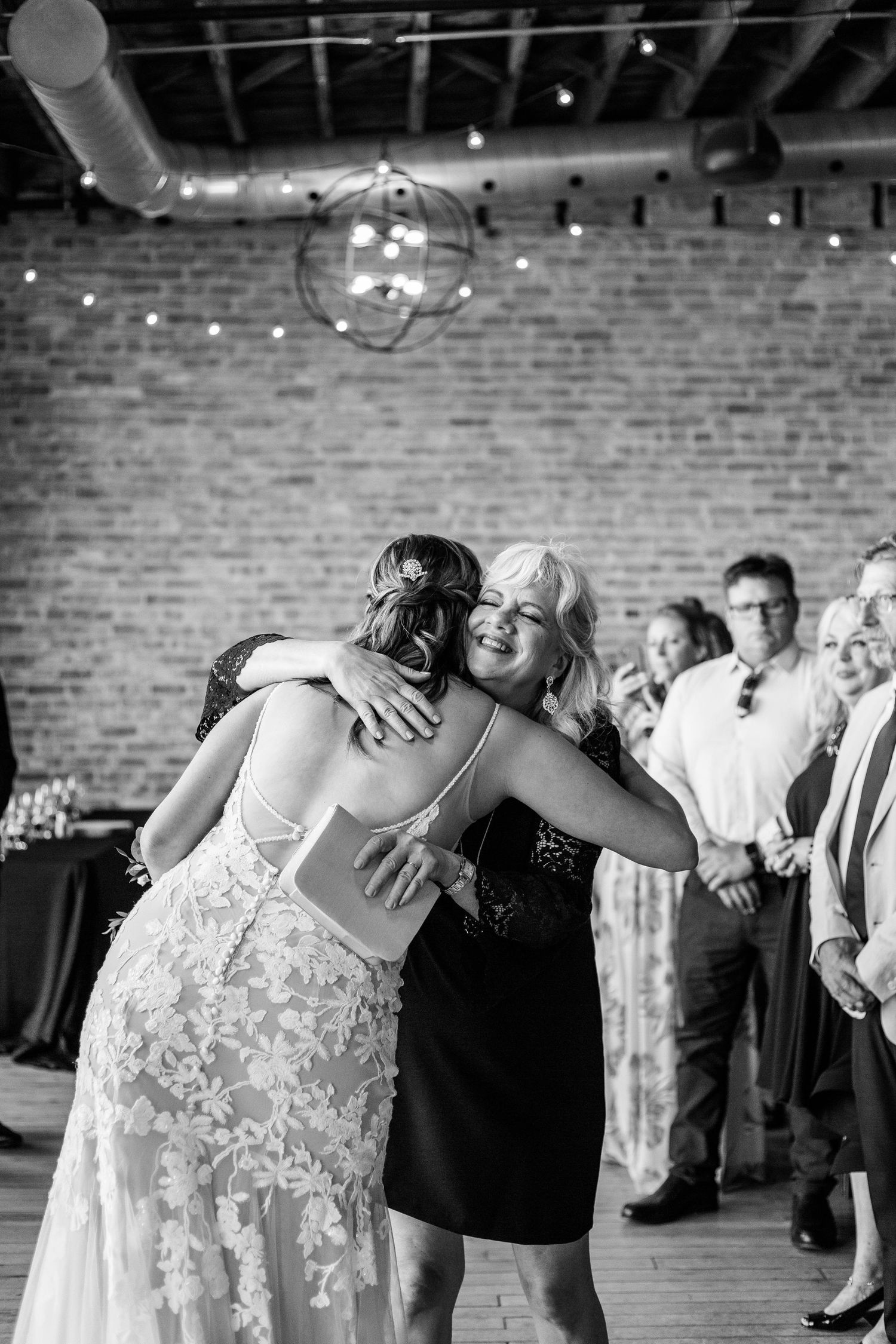 Black and white photo of bride in lace gown sharing emotional hug with mother at wedding reception.