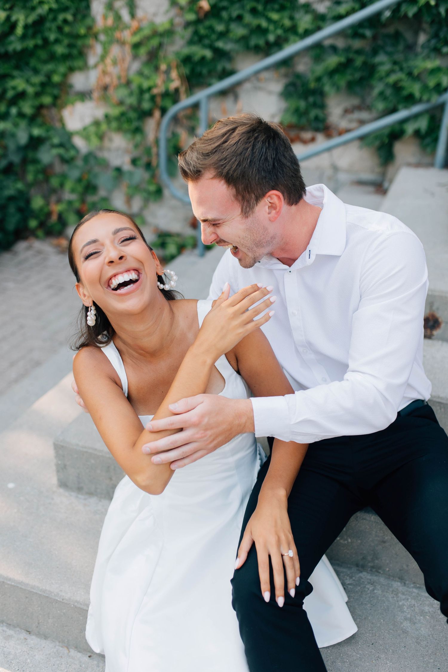 Couple laughs joyfully together on stone steps outside ivy-covered building during engagement photos.