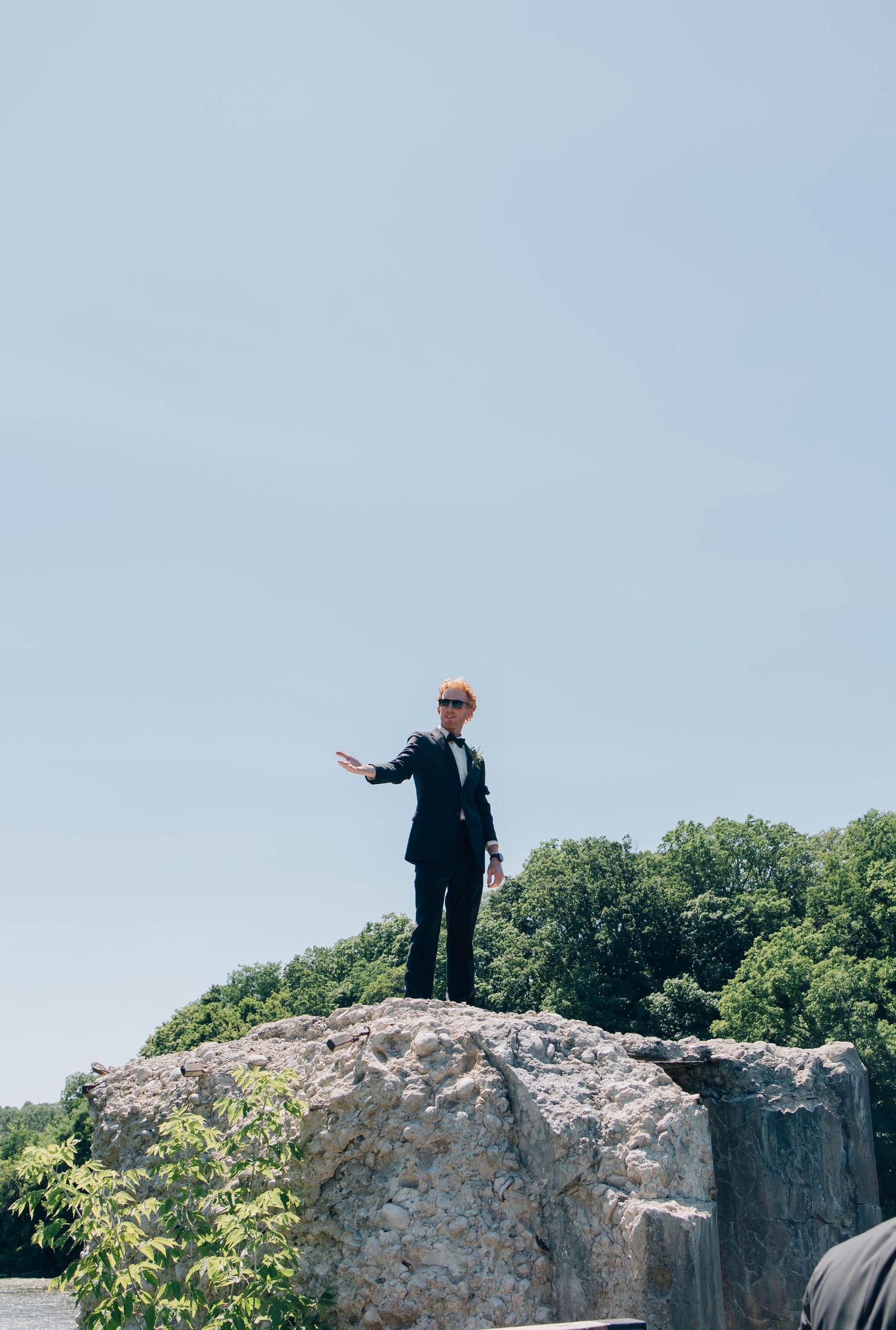 Man in black suit standing on a large rock against a clear blue sky.