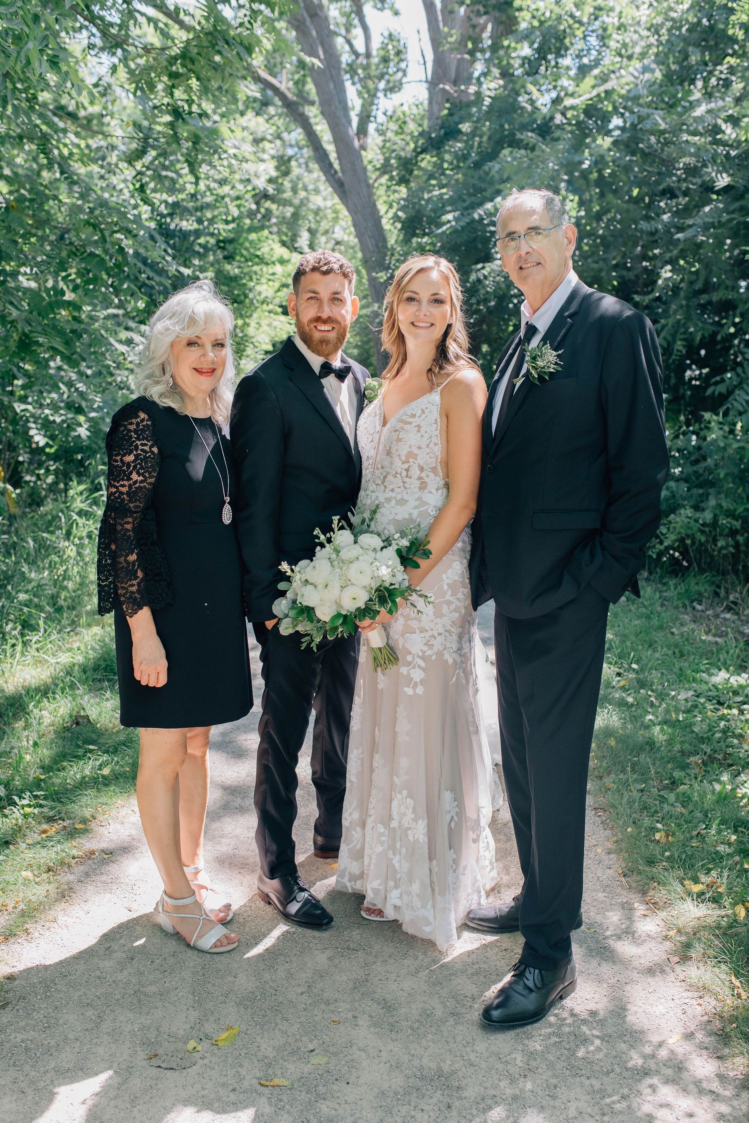 Bride and groom pose with two family members on a sunlit garden path surrounded by green trees.
