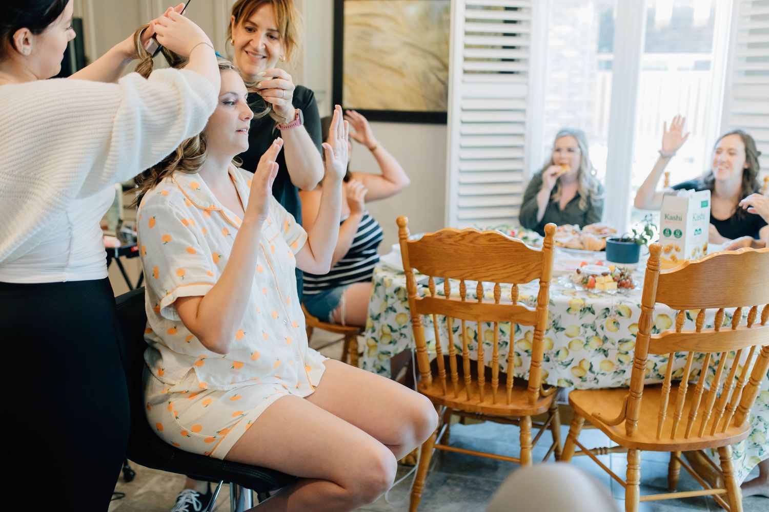 Bridesmaids laughing and taking photos during wedding morning preparations at dining room table.