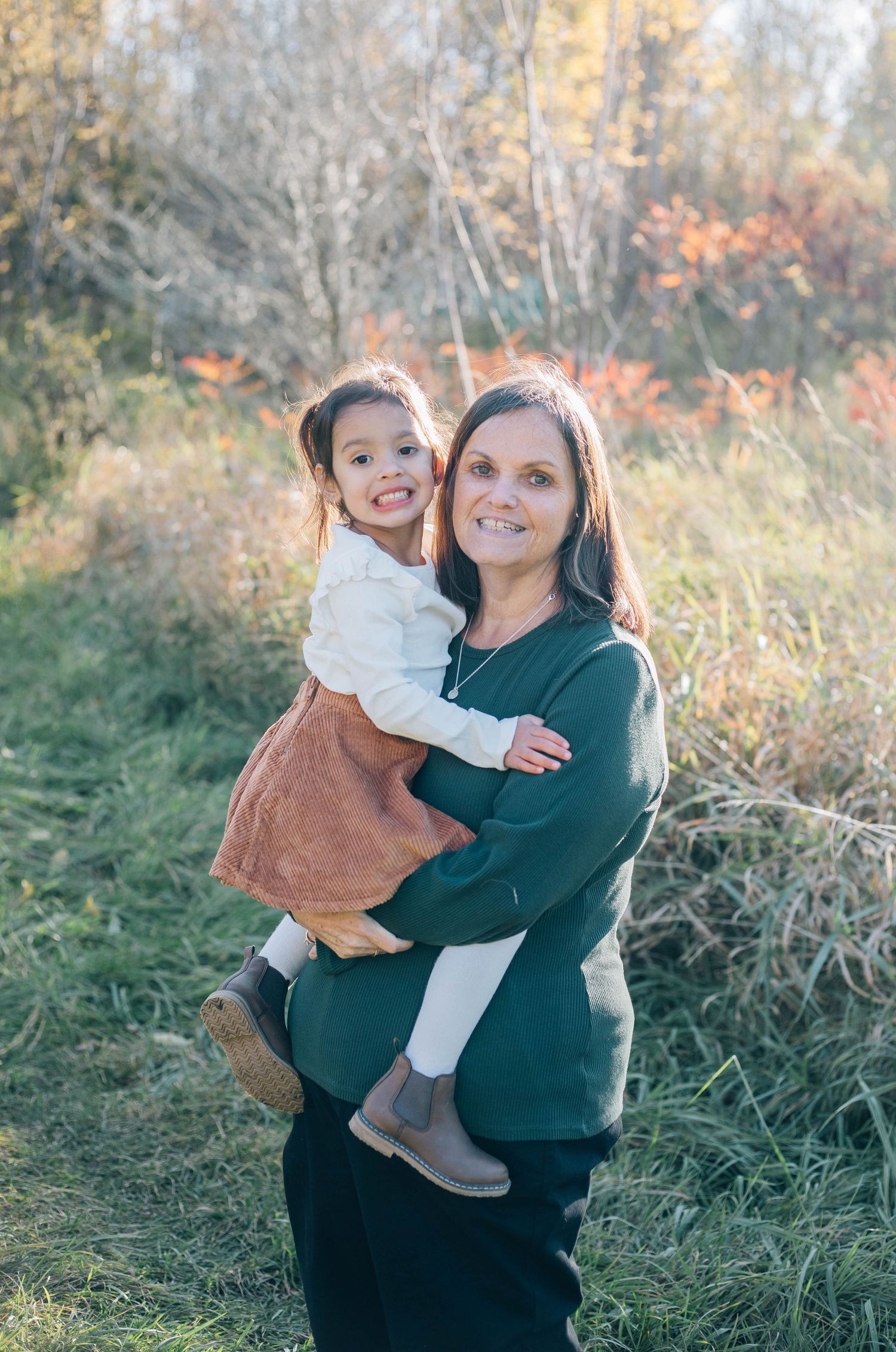 Woman in green sweater smiling while holding a young girl in rust skirt outdoors in fall foliage.