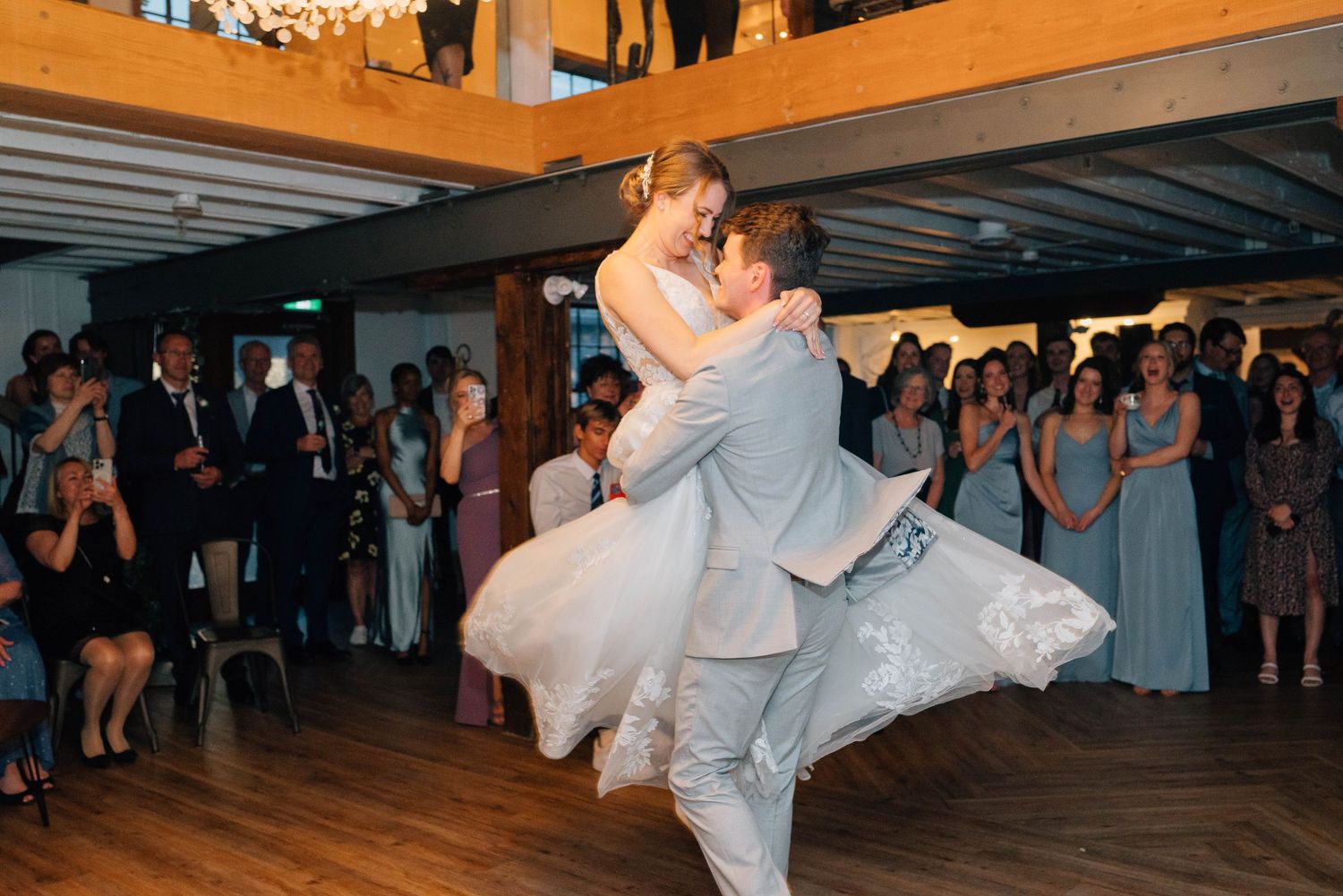 Groom lifting bride joyfully during first dance as wedding guests cheer in the background.