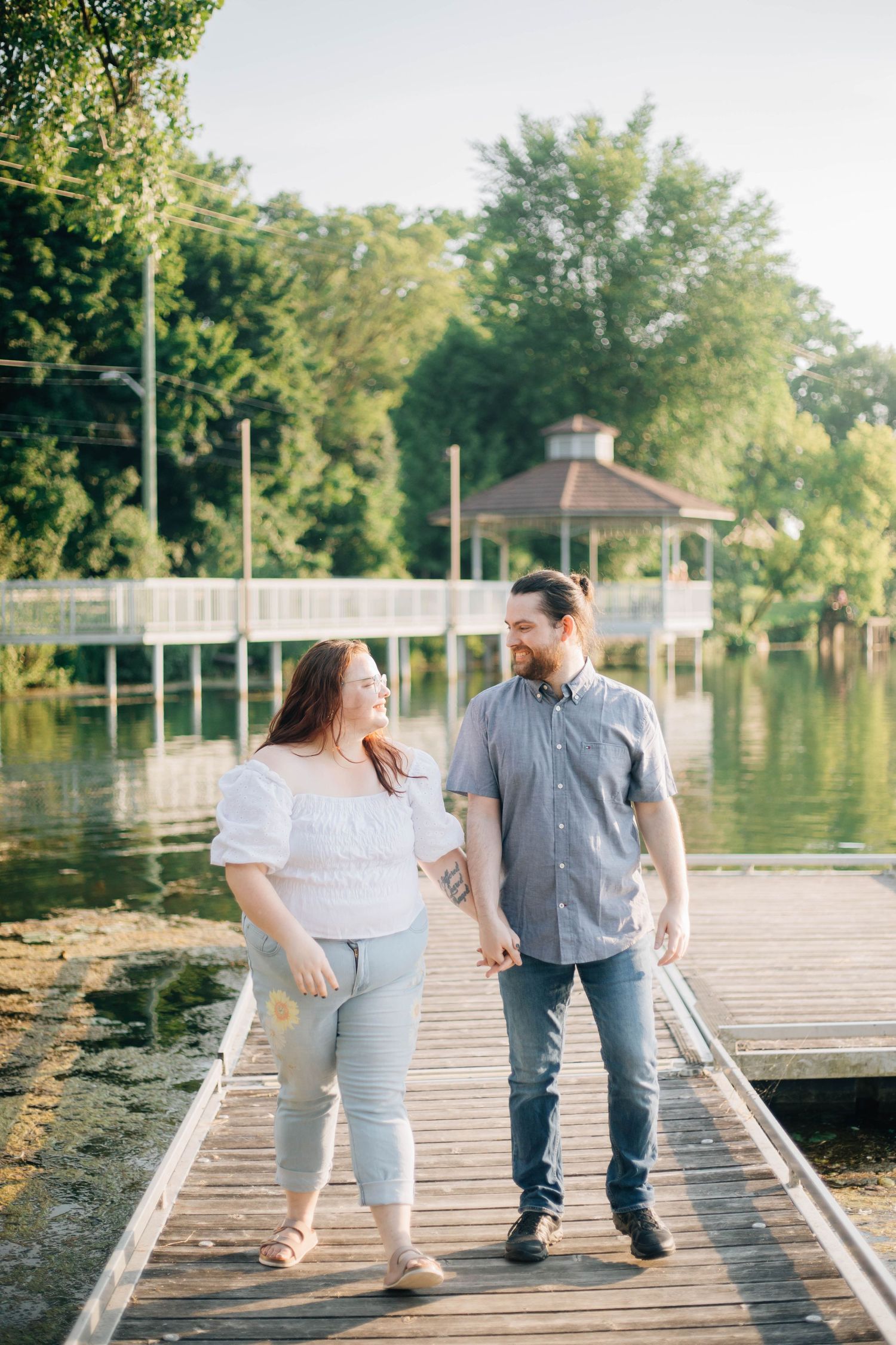 Smiling couple holding hands and walking on a wooden dock with a scenic lake and gazebo.
