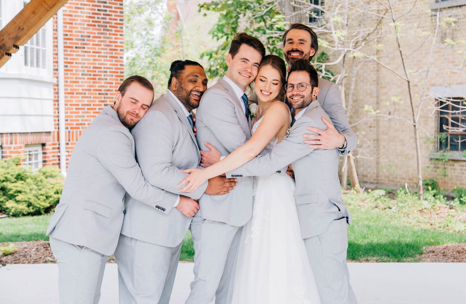 Groom surrounded by smiling groomsmen in gray suits sharing a group hug at outdoor wedding venue.