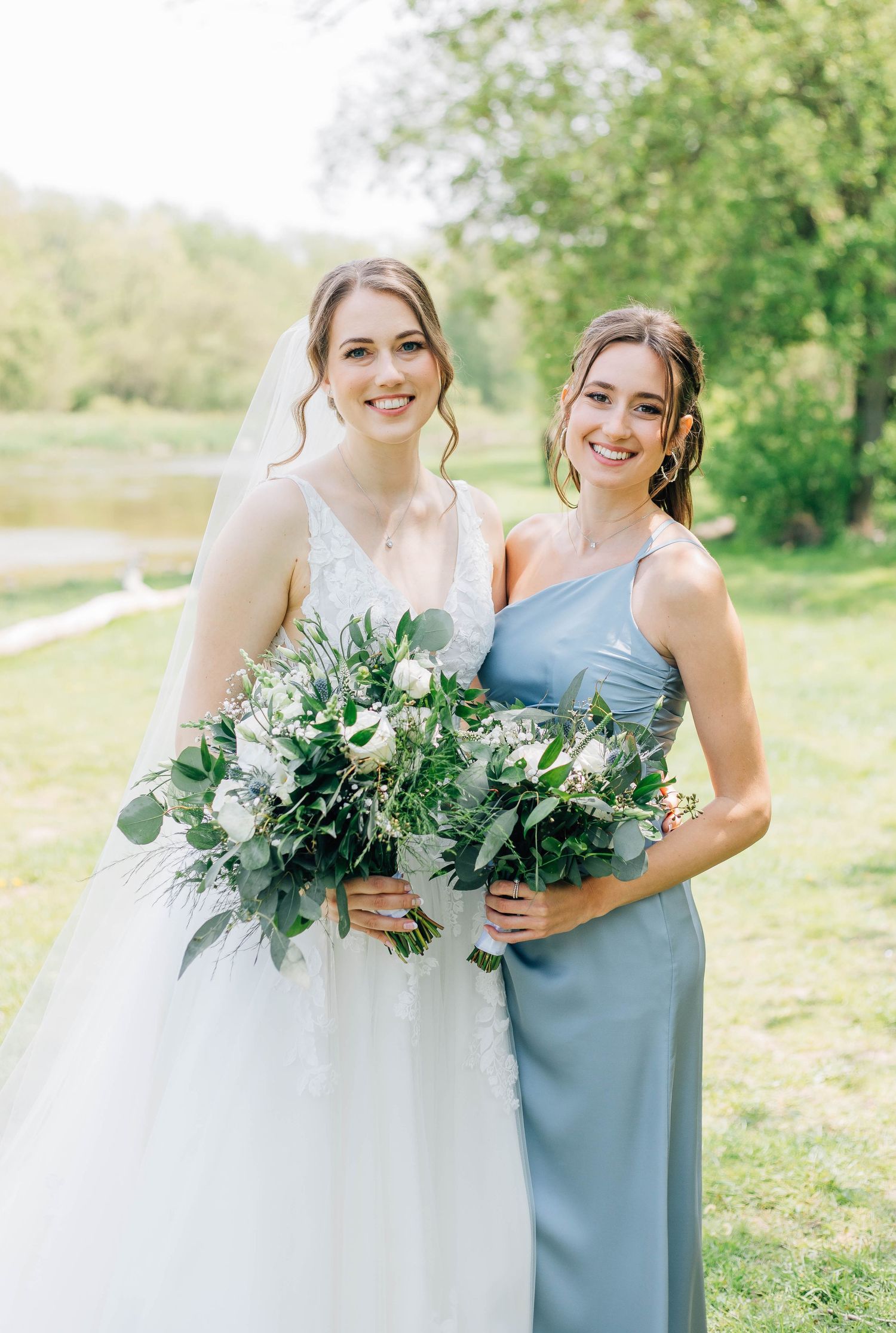 Bride in white lace gown and bridesmaid in dusty blue dress smile together outdoors by a pond.