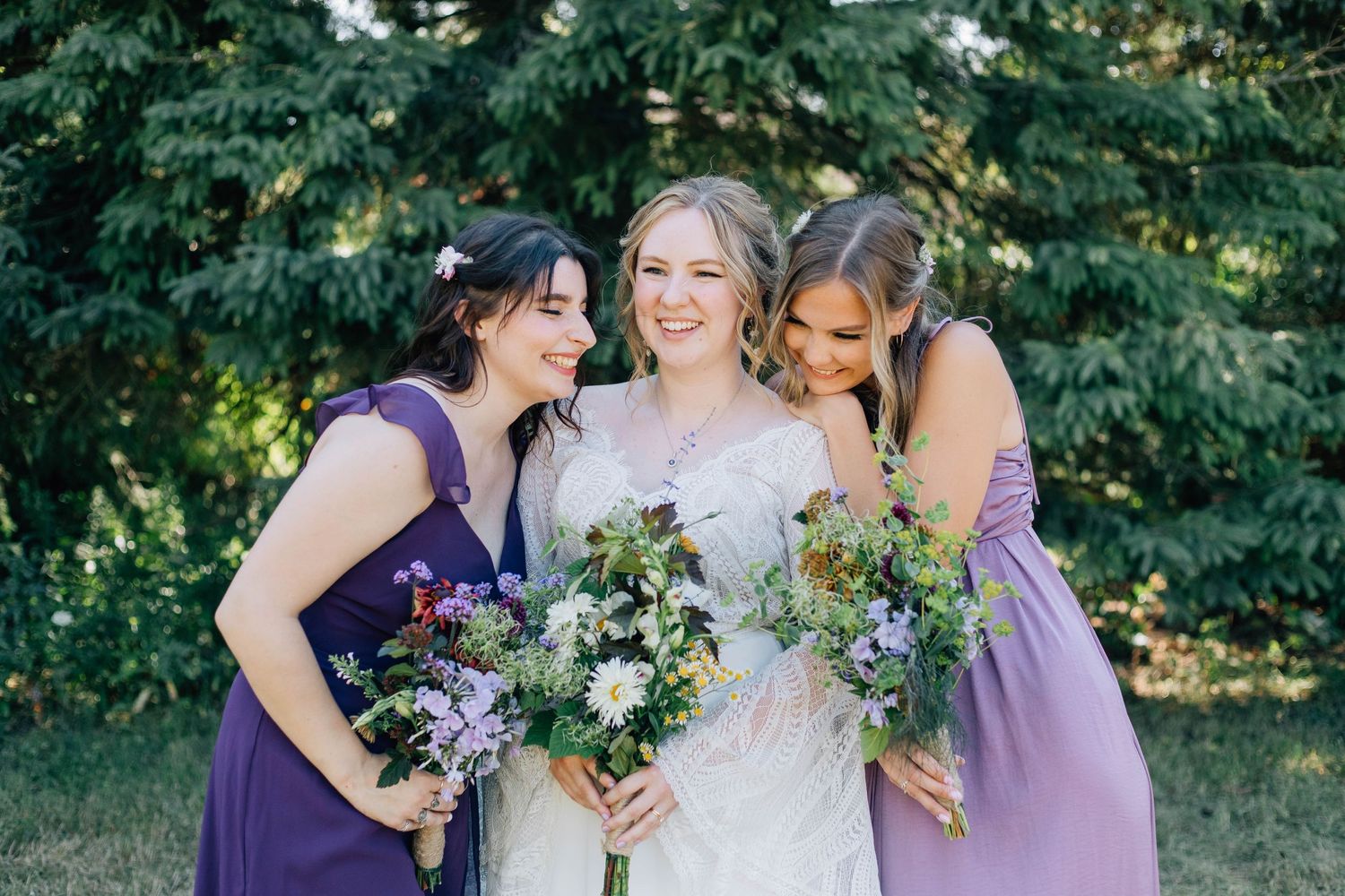 Bride smiling between two bridesmaids in purple dresses holding wildflower bouquets in a garden setting.
