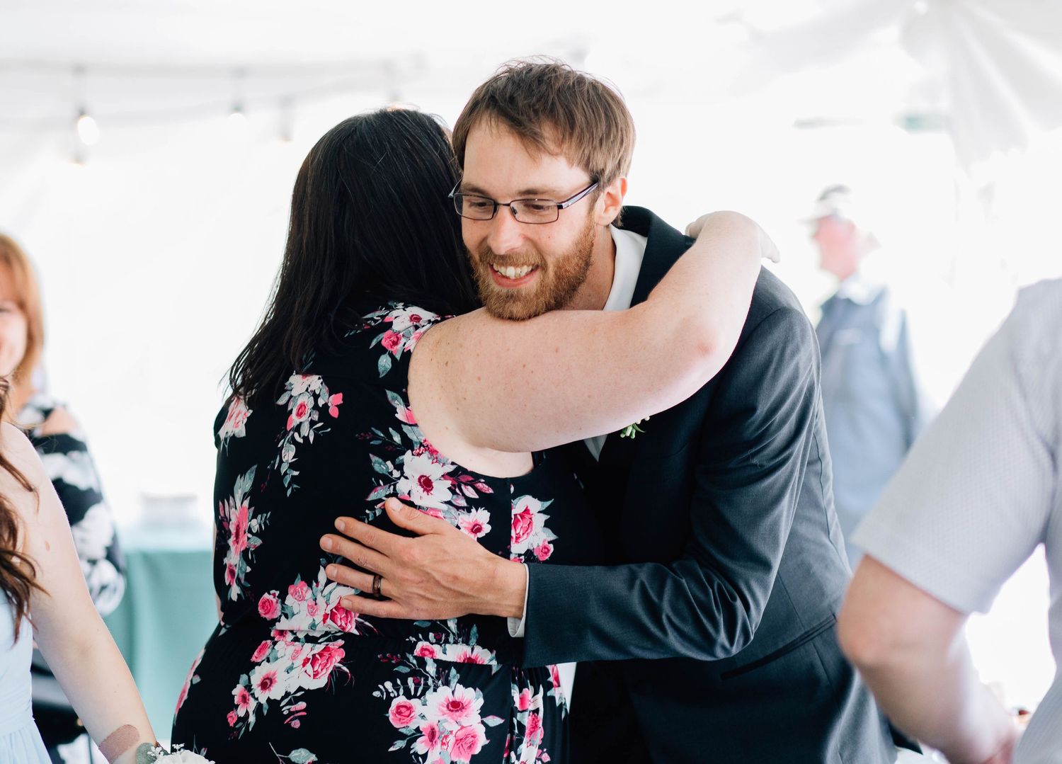 A man with glasses in a dark suit shares a warm hug with a woman in a floral dress at a wedding.