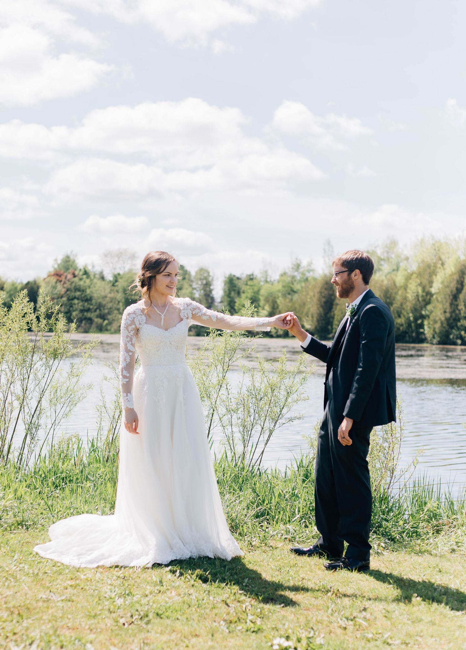 Bride and groom holding hands outdoors near a serene lake on their wedding day.