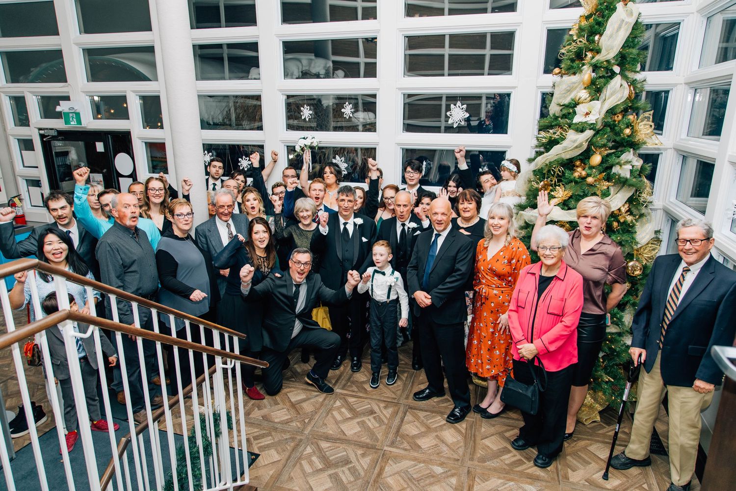 Large wedding party group photo on staircase beside decorated Christmas tree at festive winter wedding.