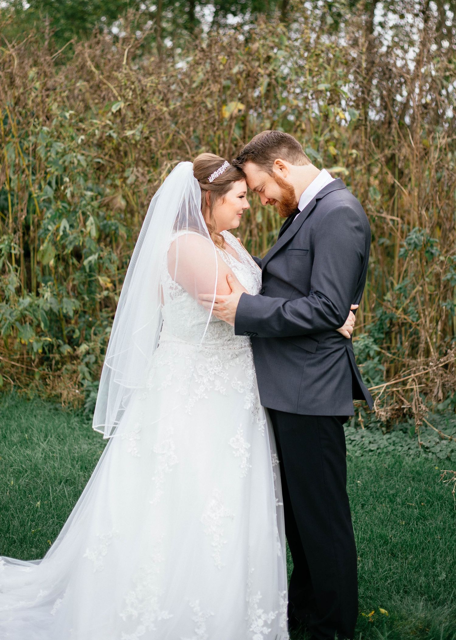 Bride and groom sharing tender forehead-to-forehead moment in lace gown amid autumn foliage.