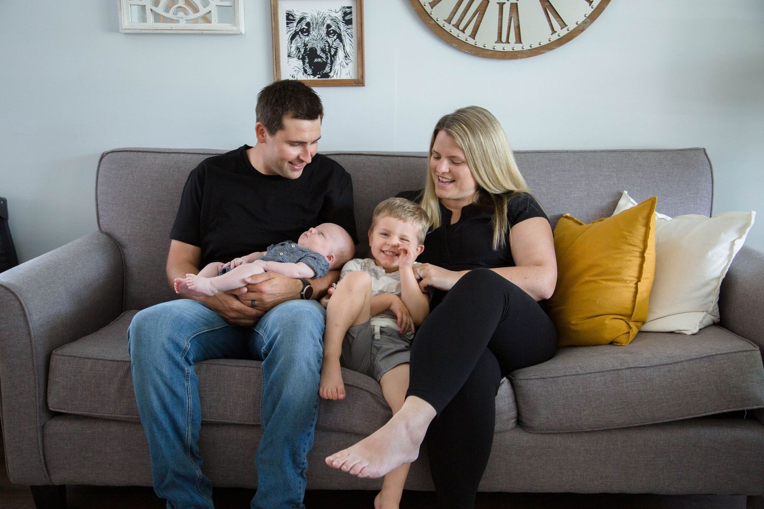 Happy family of four with newborn baby and toddler sitting together on gray couch at home.