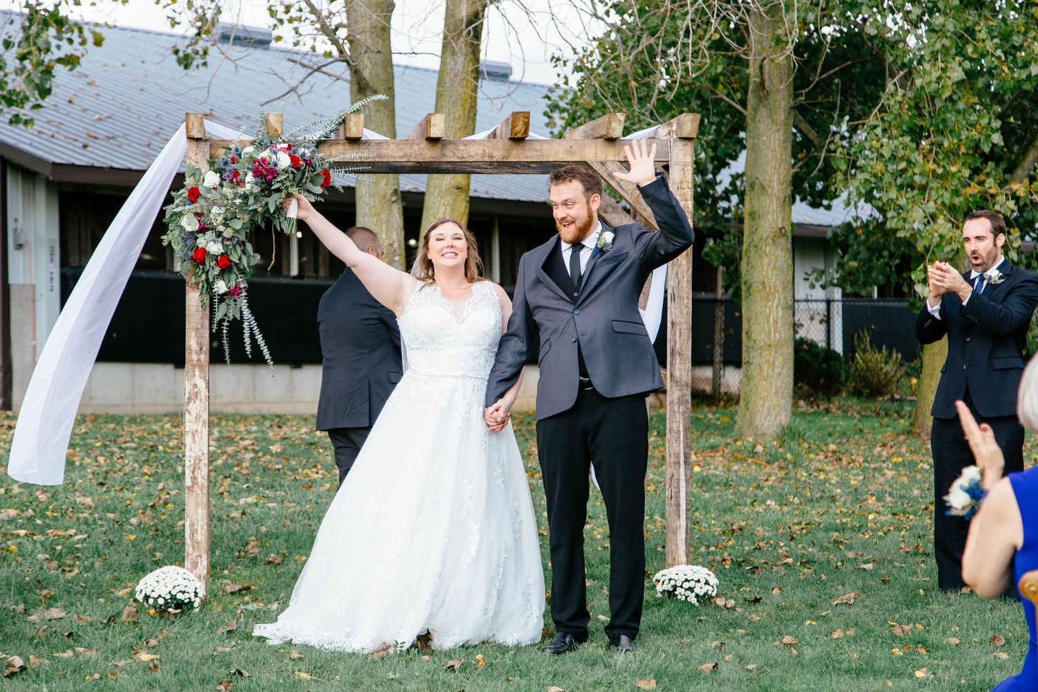Joyful newlyweds celebrate with raised arms after outdoor wedding ceremony beneath rustic wooden arbor.