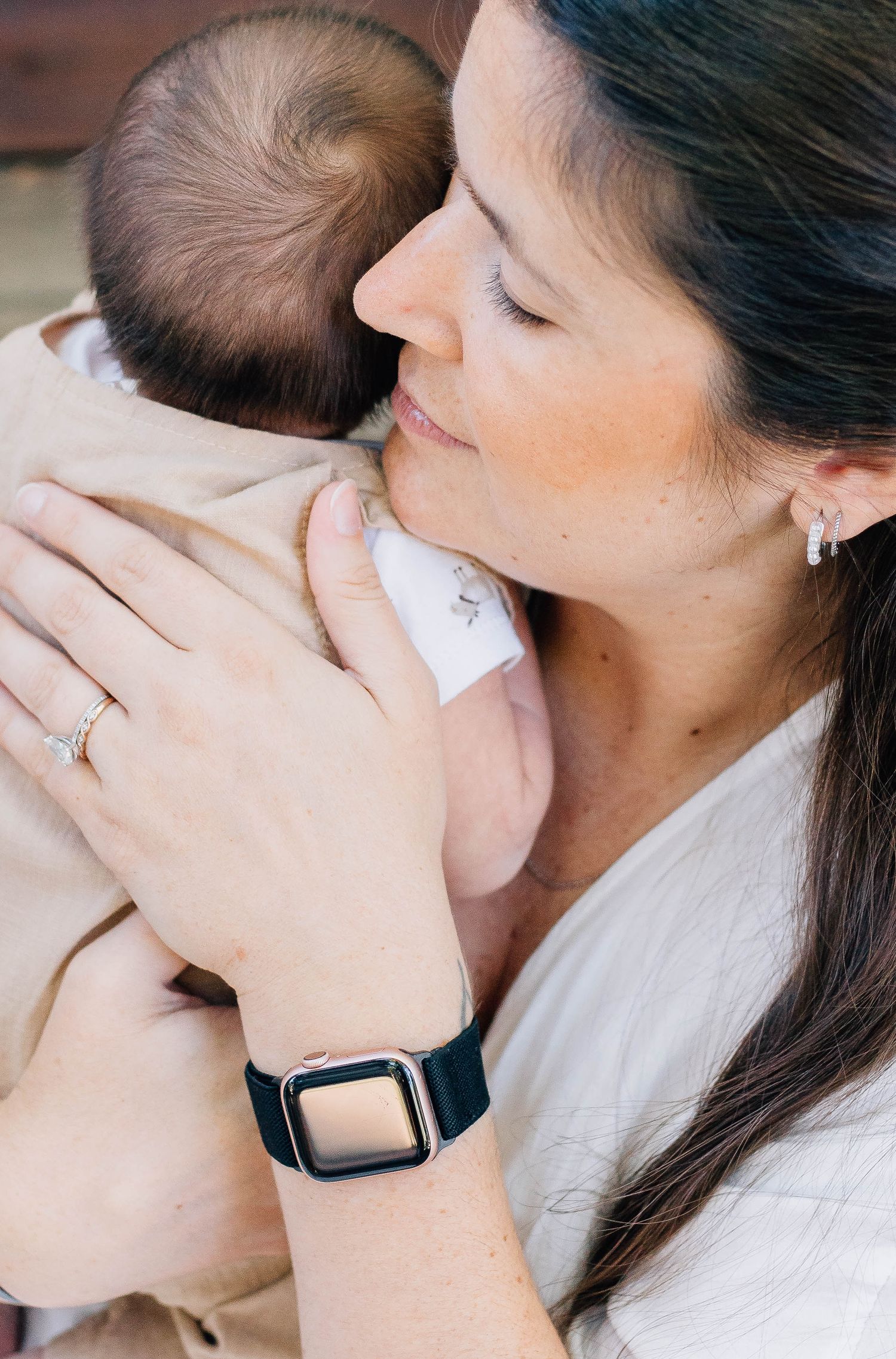 Tender close-up of dark-haired mother wearing Apple Watch and diamond ring gently nuzzling her sleeping newborn baby.