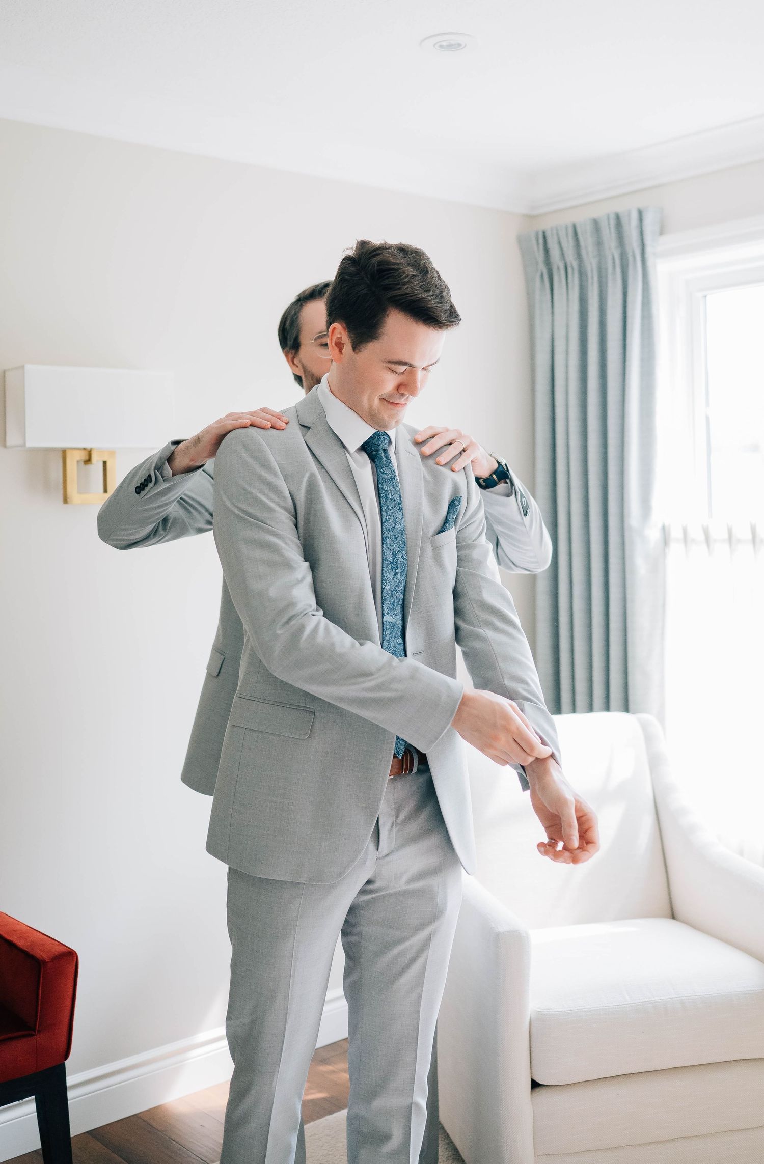 Groom in light gray suit smiles while adjusting his cufflinks with a groomsman nearby.