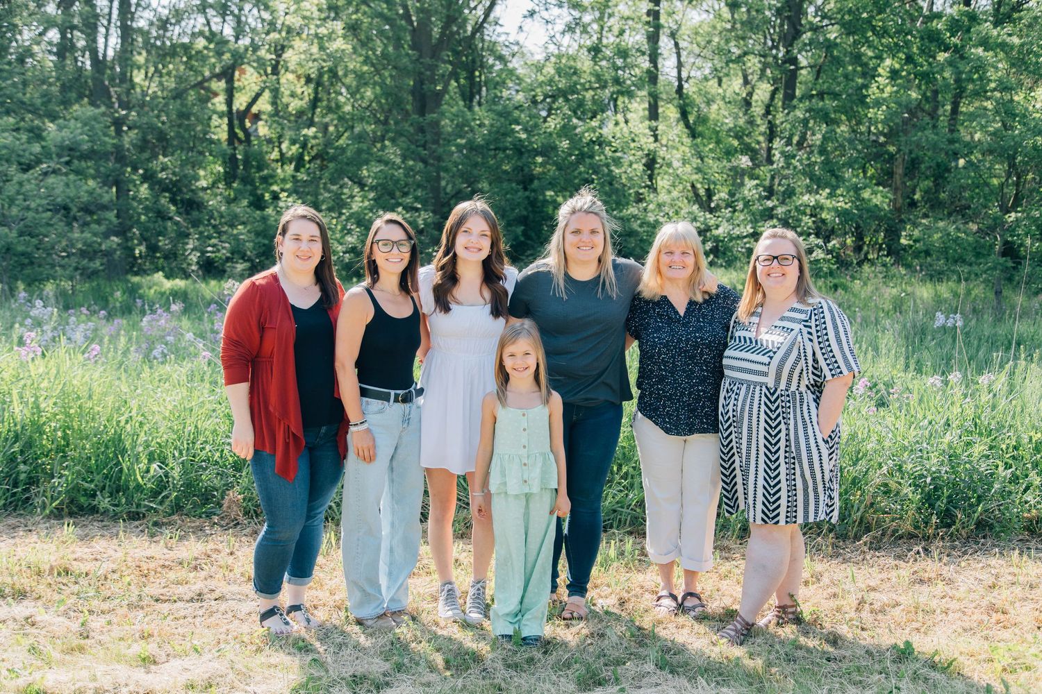 Seven women and girls smiling together outdoors in a sunny green field for a family portrait.