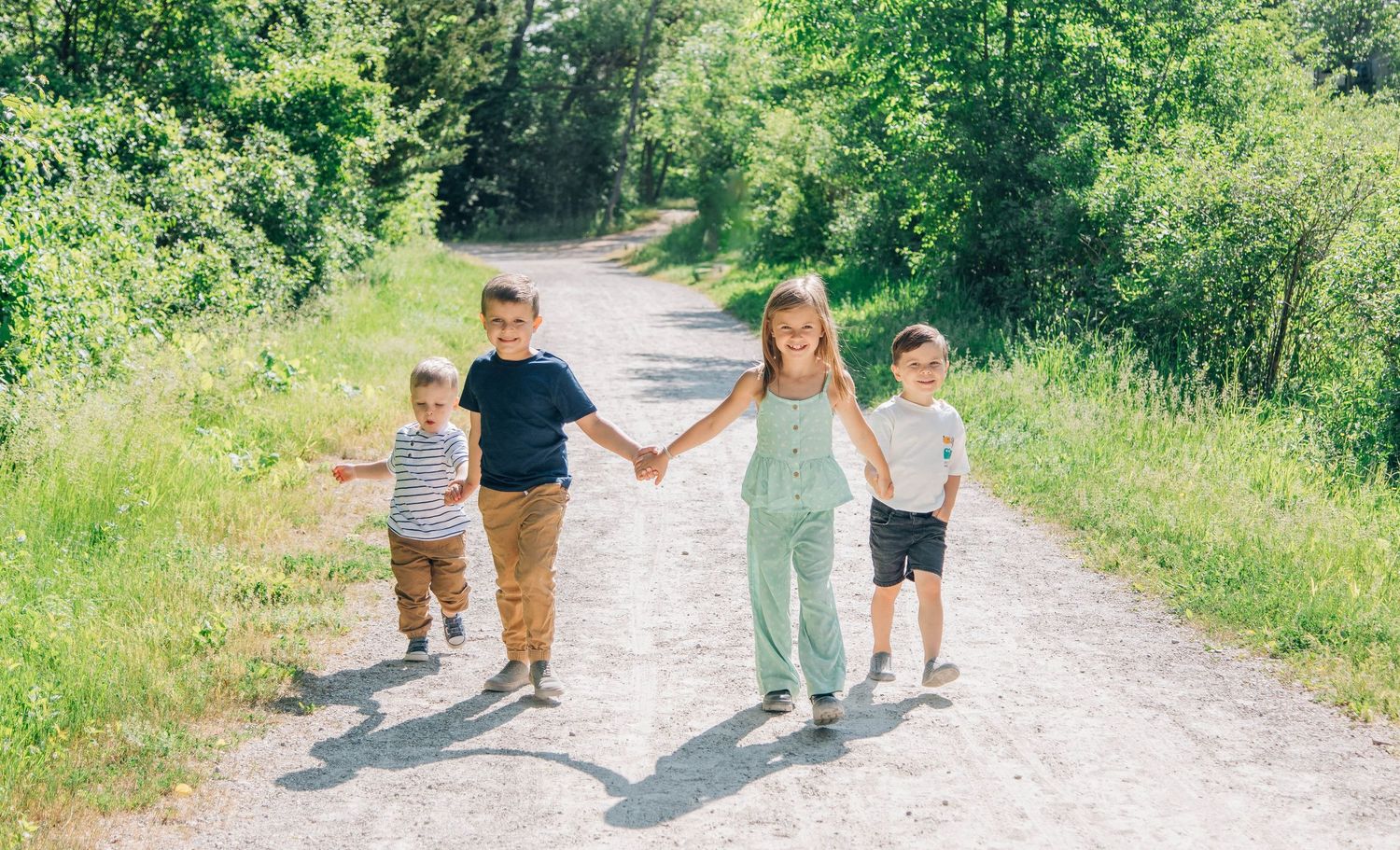 Four young children holding hands and walking together on a sunny tree-lined path.