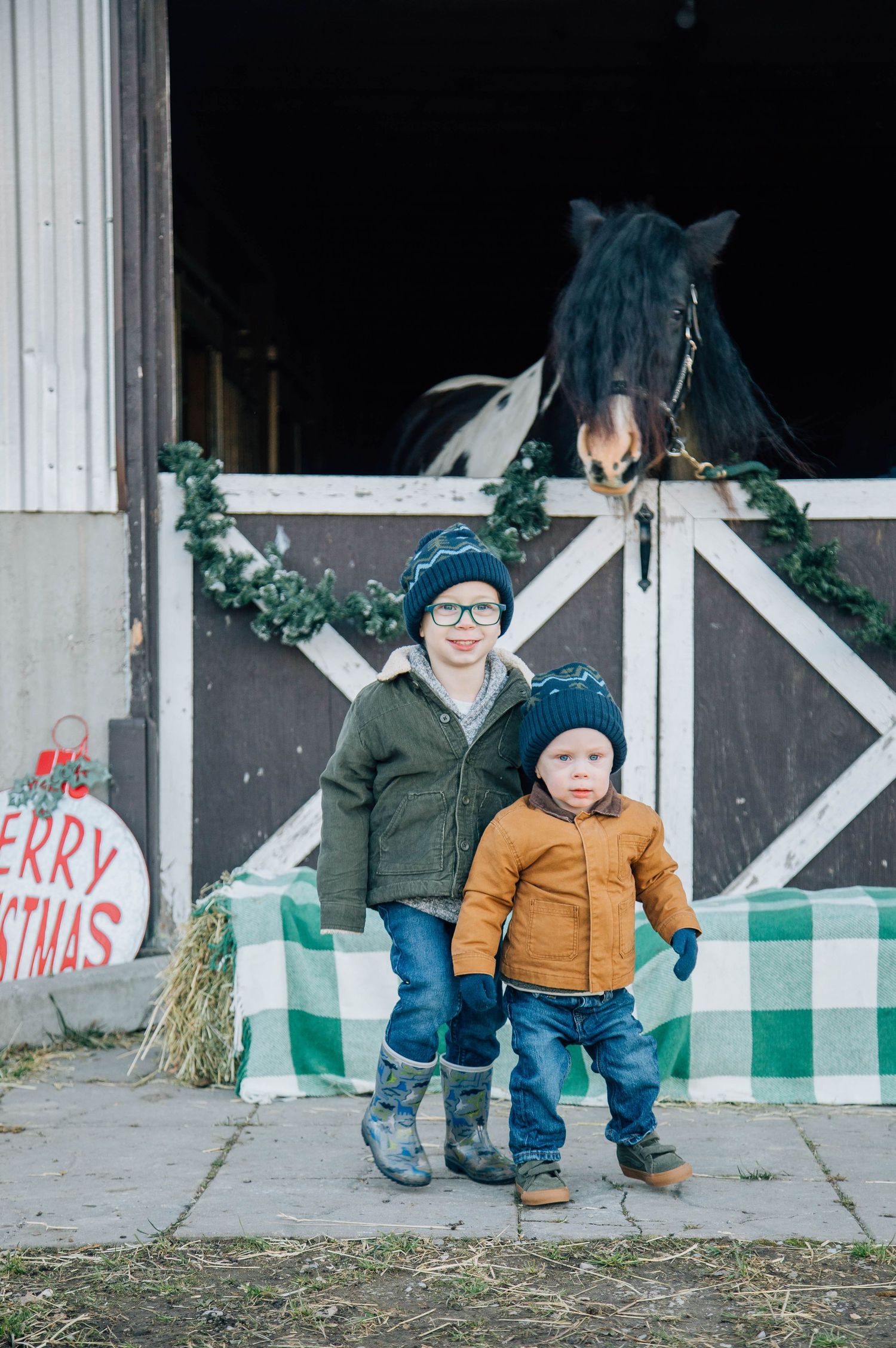 Two young boys pose in front of a decorated horse stable with a horse peering over the door at a Christmas farm setting.