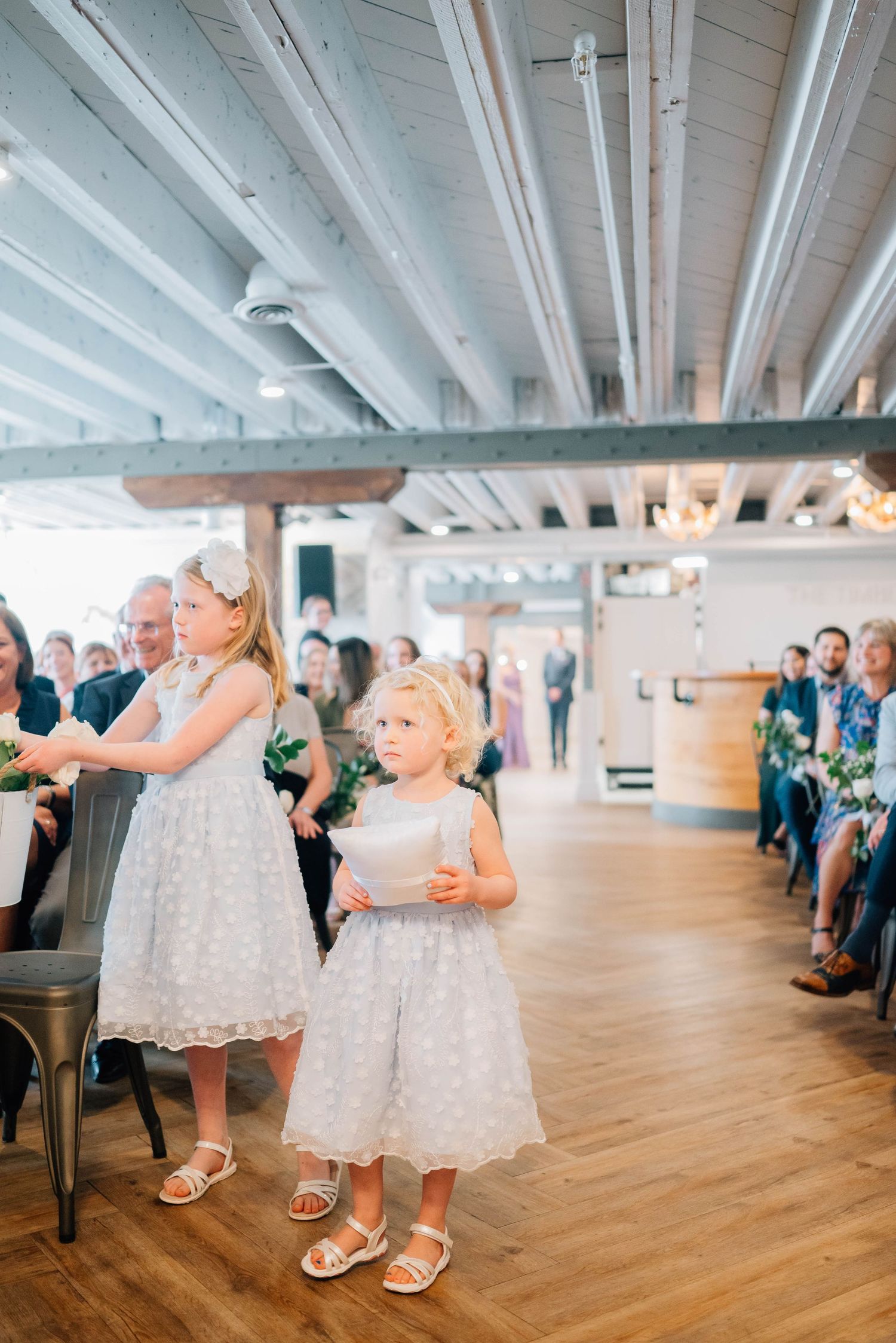 Two flower girls in white lace dresses walking down wedding ceremony aisle.