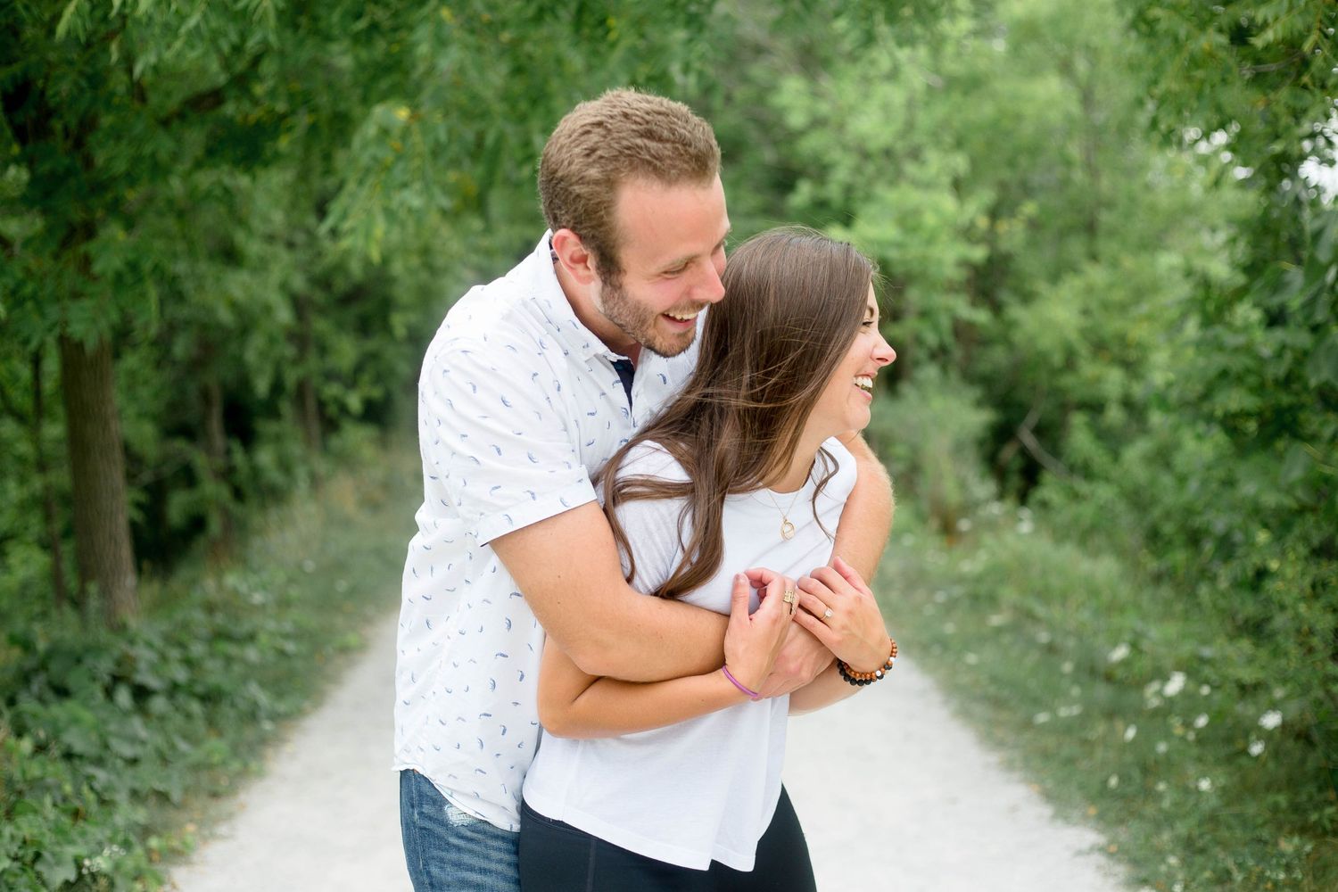 Couple embracing and laughing together on a tree-lined gravel path during an outdoor engagement photo session.