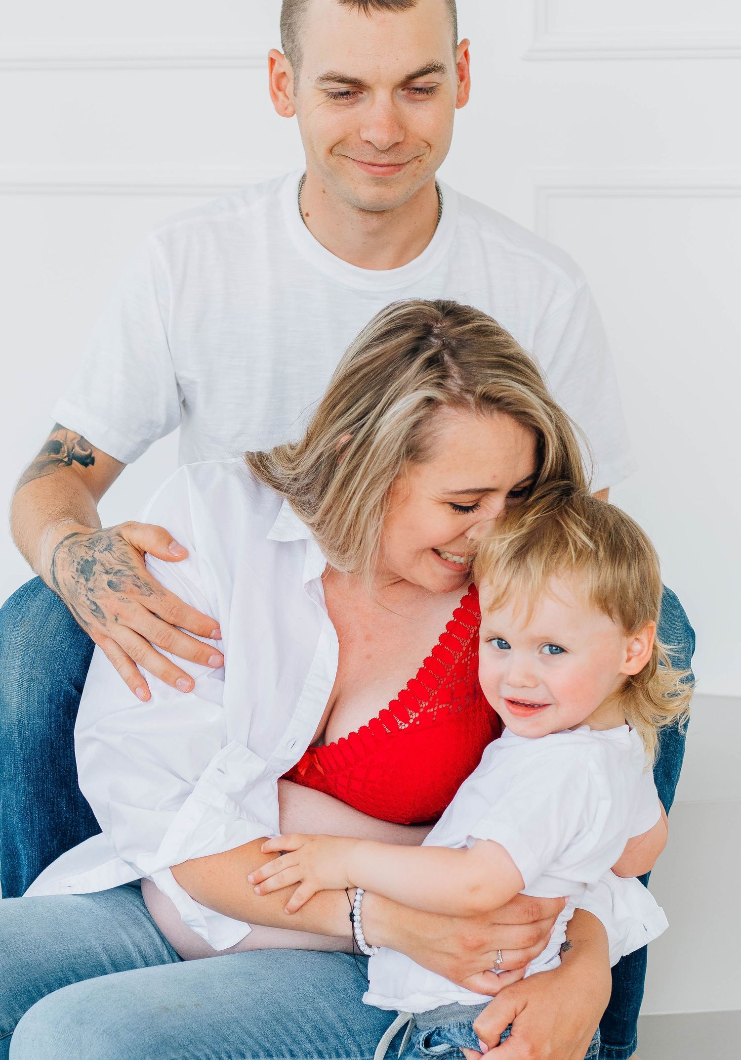 Tender family maternity moment with tattooed dad embracing pregnant blonde mom holding smiling toddler in white studio.