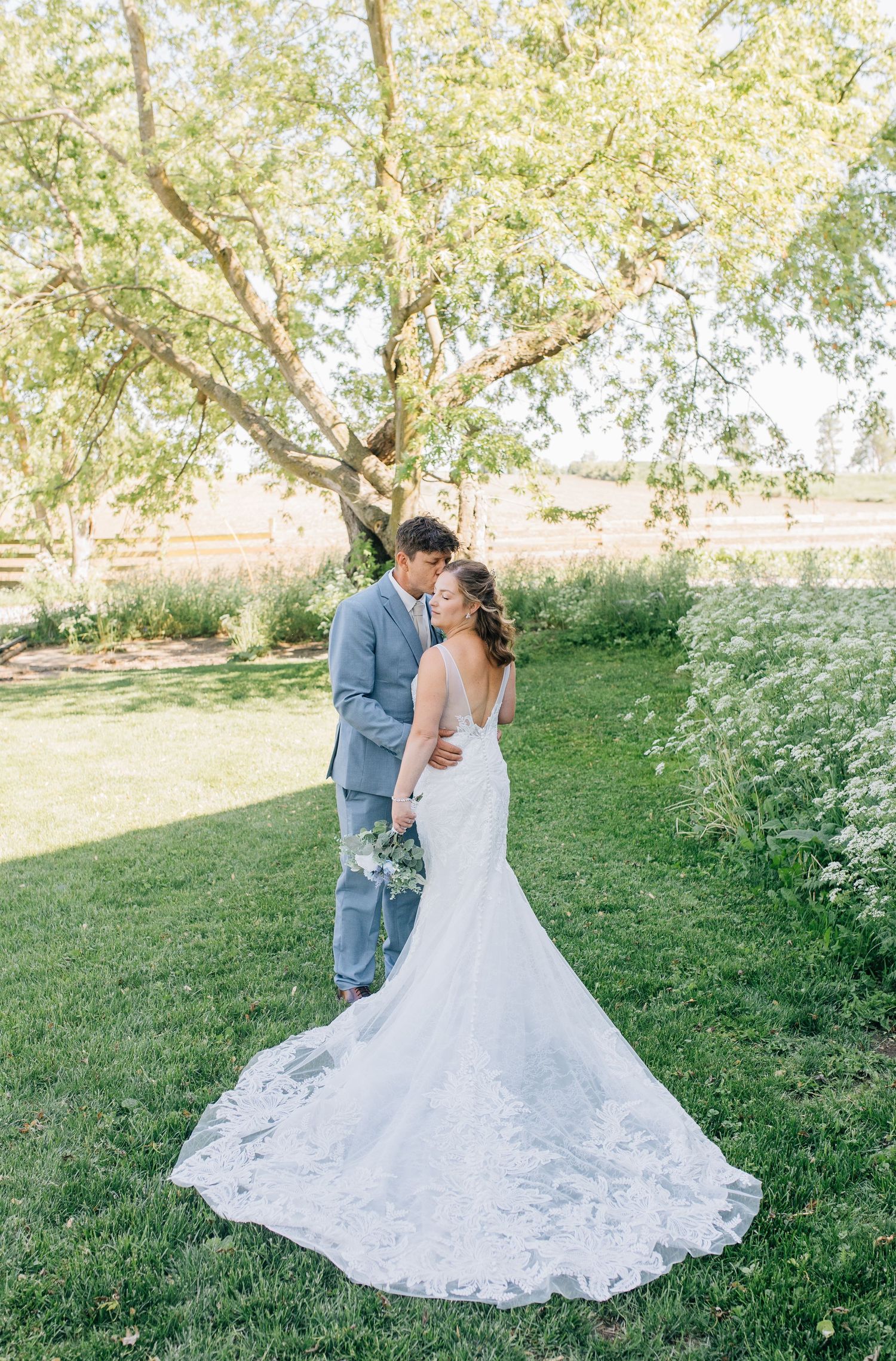 Bride in lace gown with train and groom in gray suit share a romantic moment on green lawn under large tree at venue in Woodstock Ontario